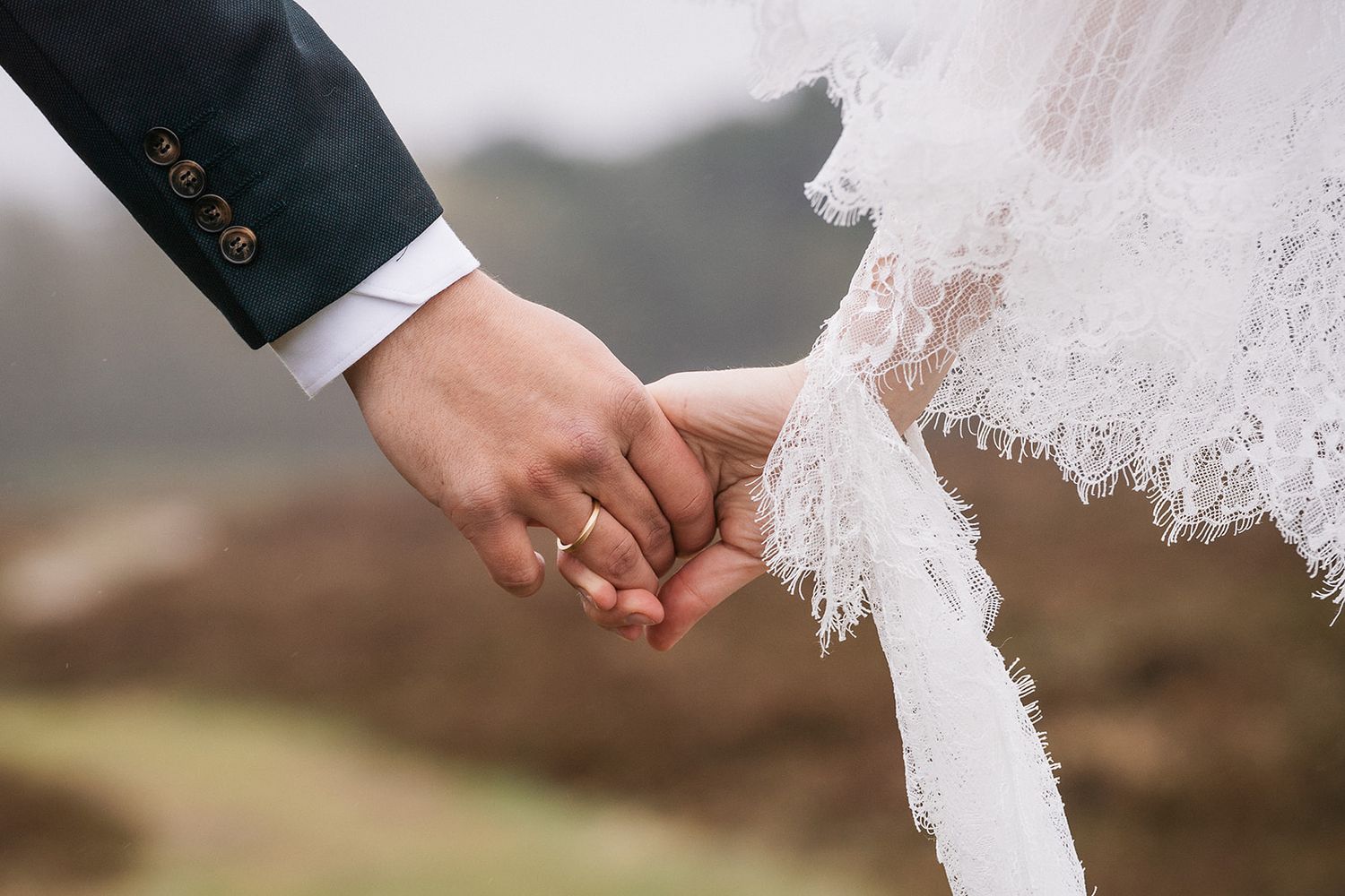 A close-up shot of newlywed couple holding hands, with lace wedding dress detail and suit sleeve visible.