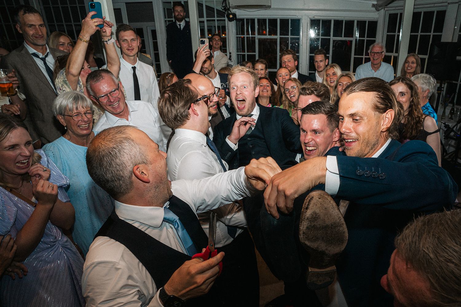 Lively party crowd celebrating and dancing at an indoor event with visible windows in background.