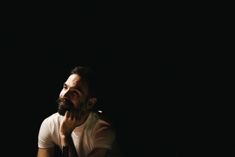Person in white shirt sitting contemplatively against dark background with dramatic side lighting.