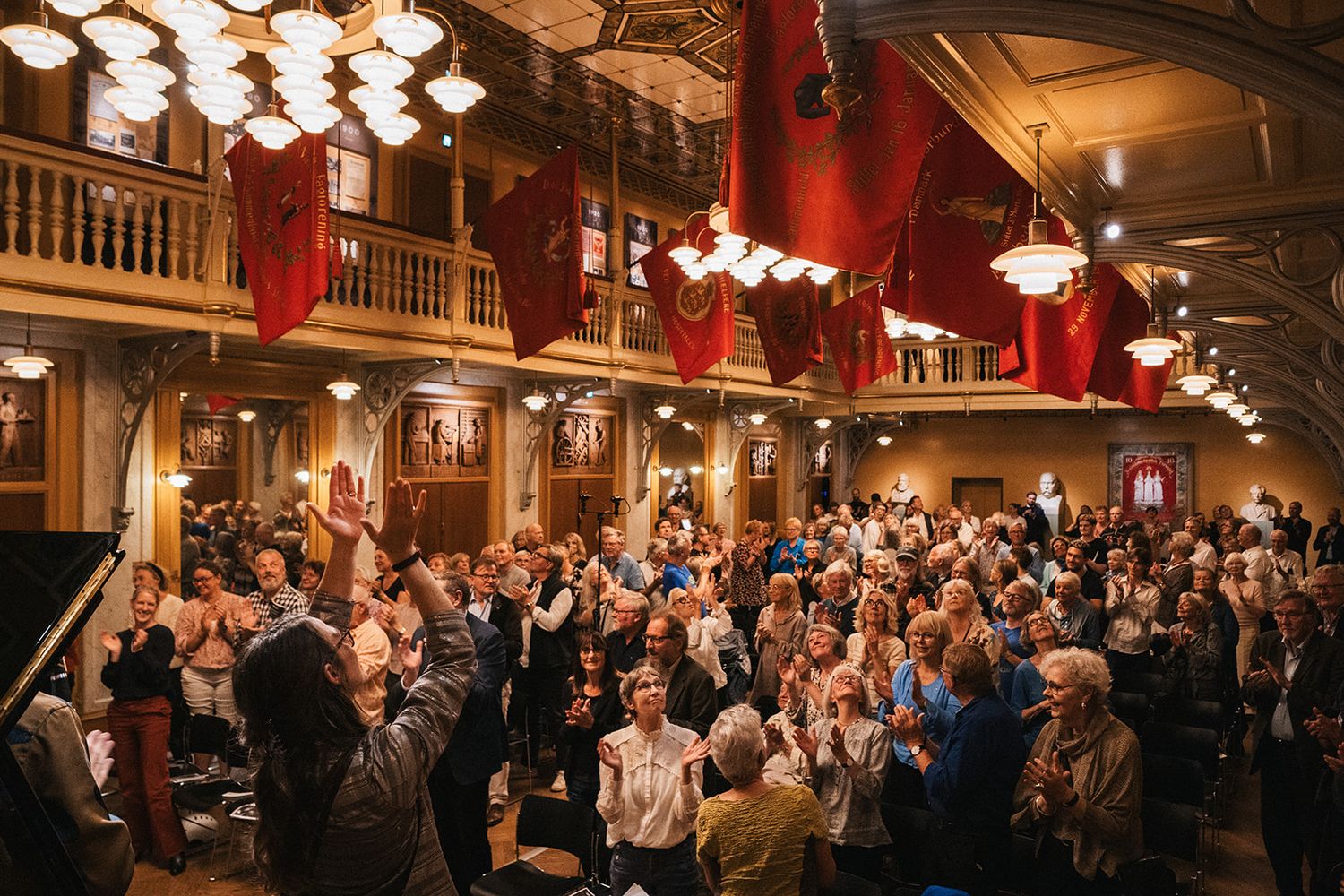 A crowded historic theater venue with ornate wooden balconies, red decor and vintage chandeliers filled with standing attendees.