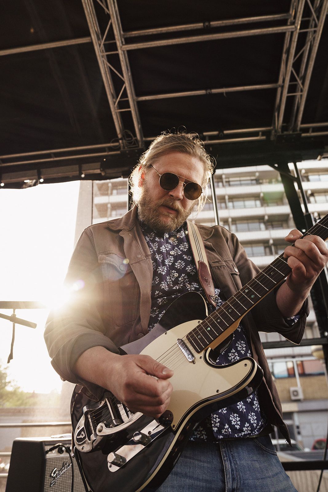 A musician performs on stage with an electric guitar at sunset wearing sunglasses and a floral print shirt.