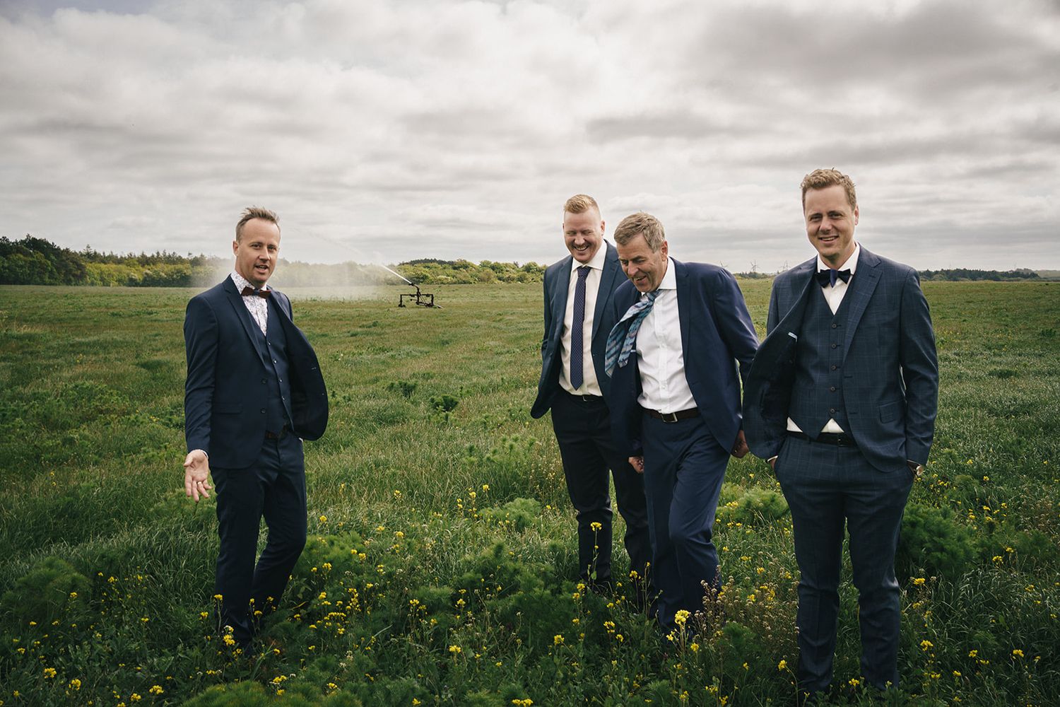 A group poses together in formal wear on a green field under cloudy skies.