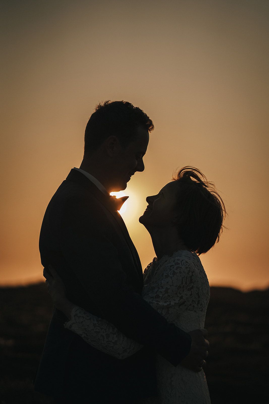 Silhouetted couple shares an intimate moment against a golden sunset sky, casting romantic shadows in a wedding photo.