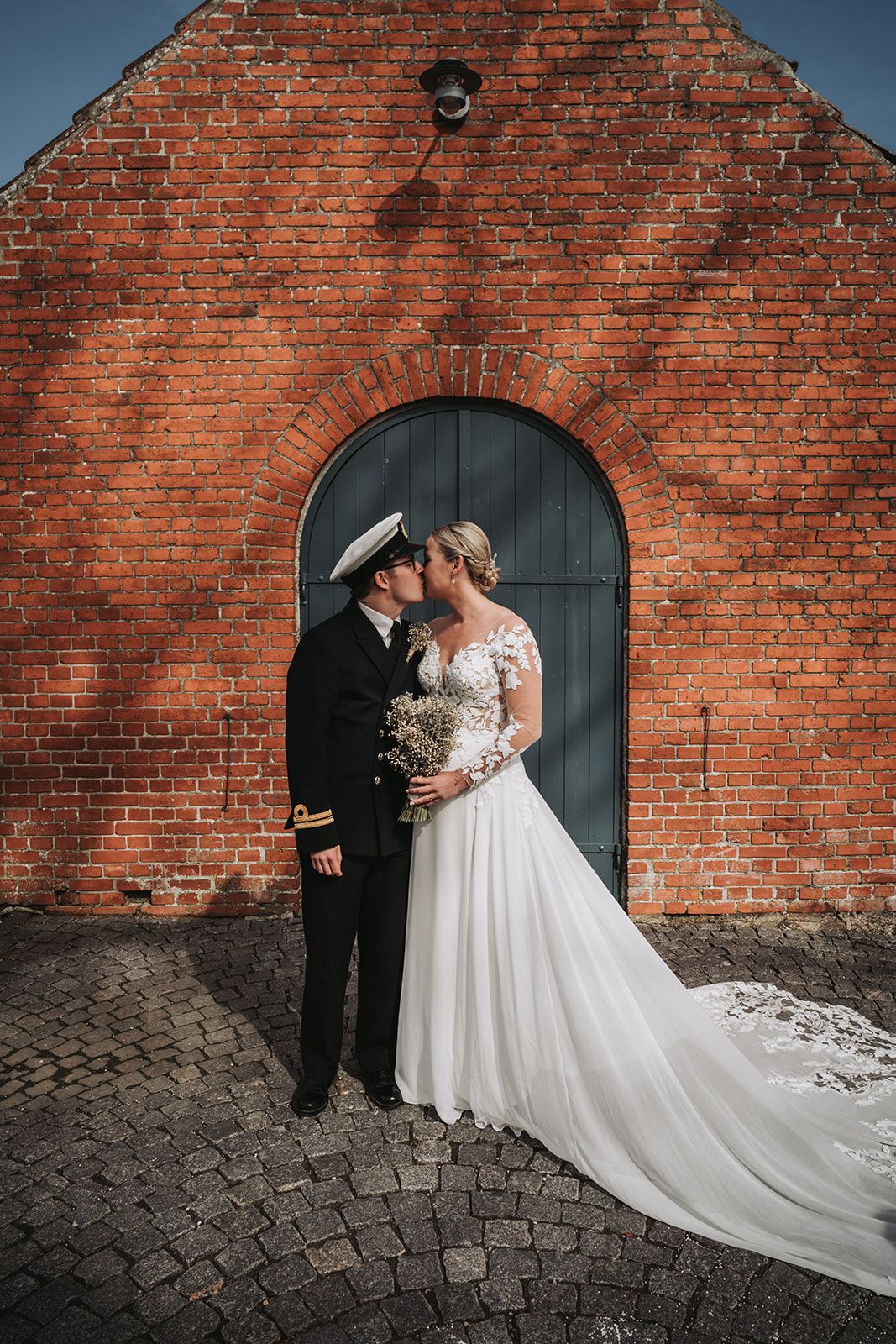A romantic wedding couple poses against a rustic red brick wall with an arched doorway and cobblestone ground.