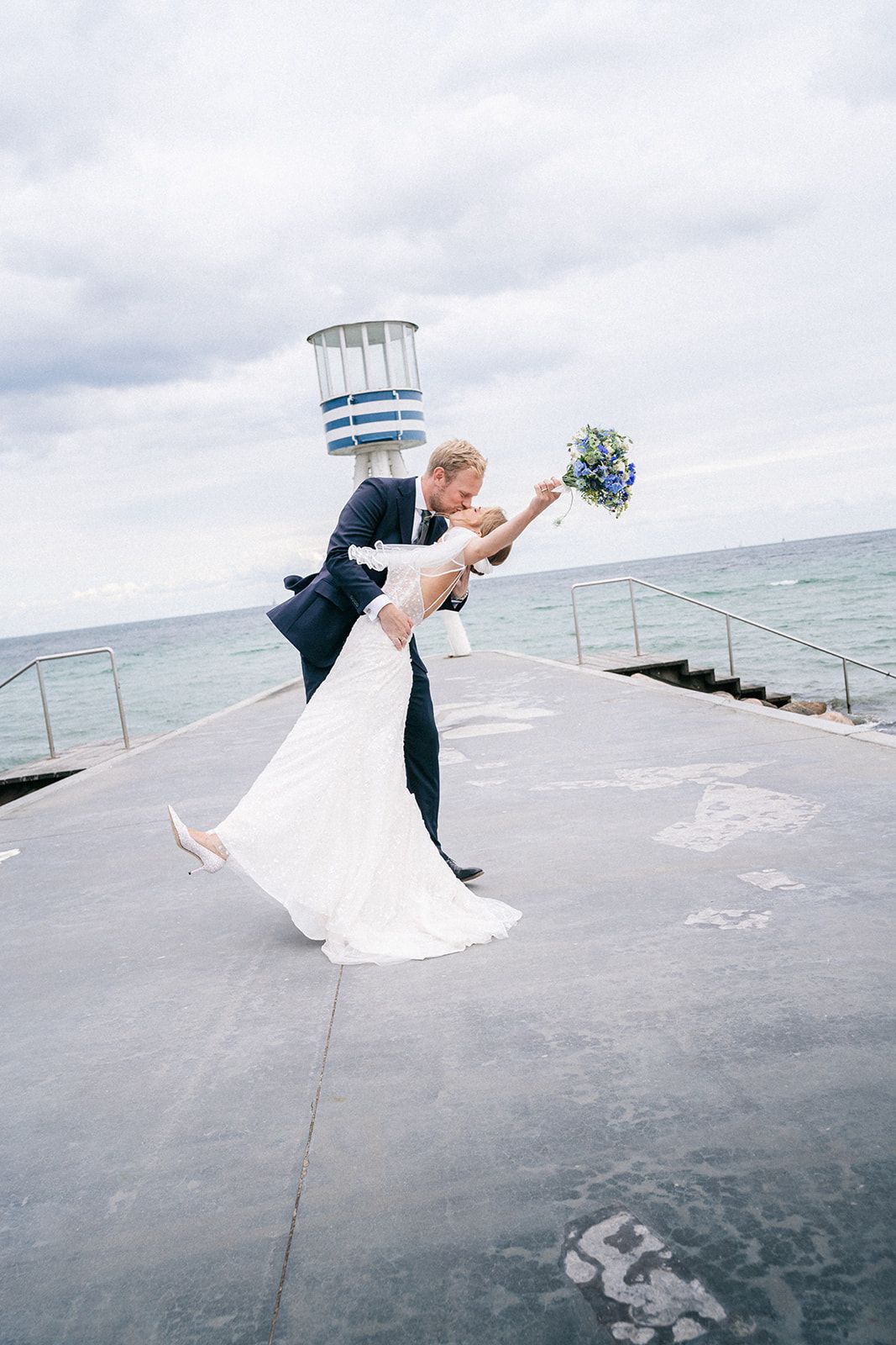 A wedding couple dips dramatically on a concrete pier overlooking the ocean with a lighthouse tower in the background.