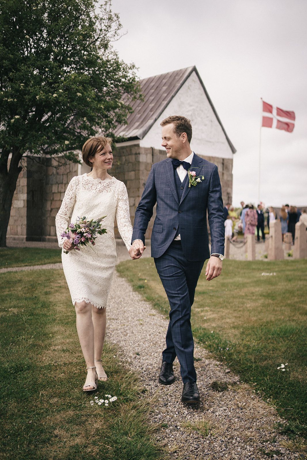 Bride in lace dress and groom in navy suit walk on gravel path near rustic building with Danish flag flying.