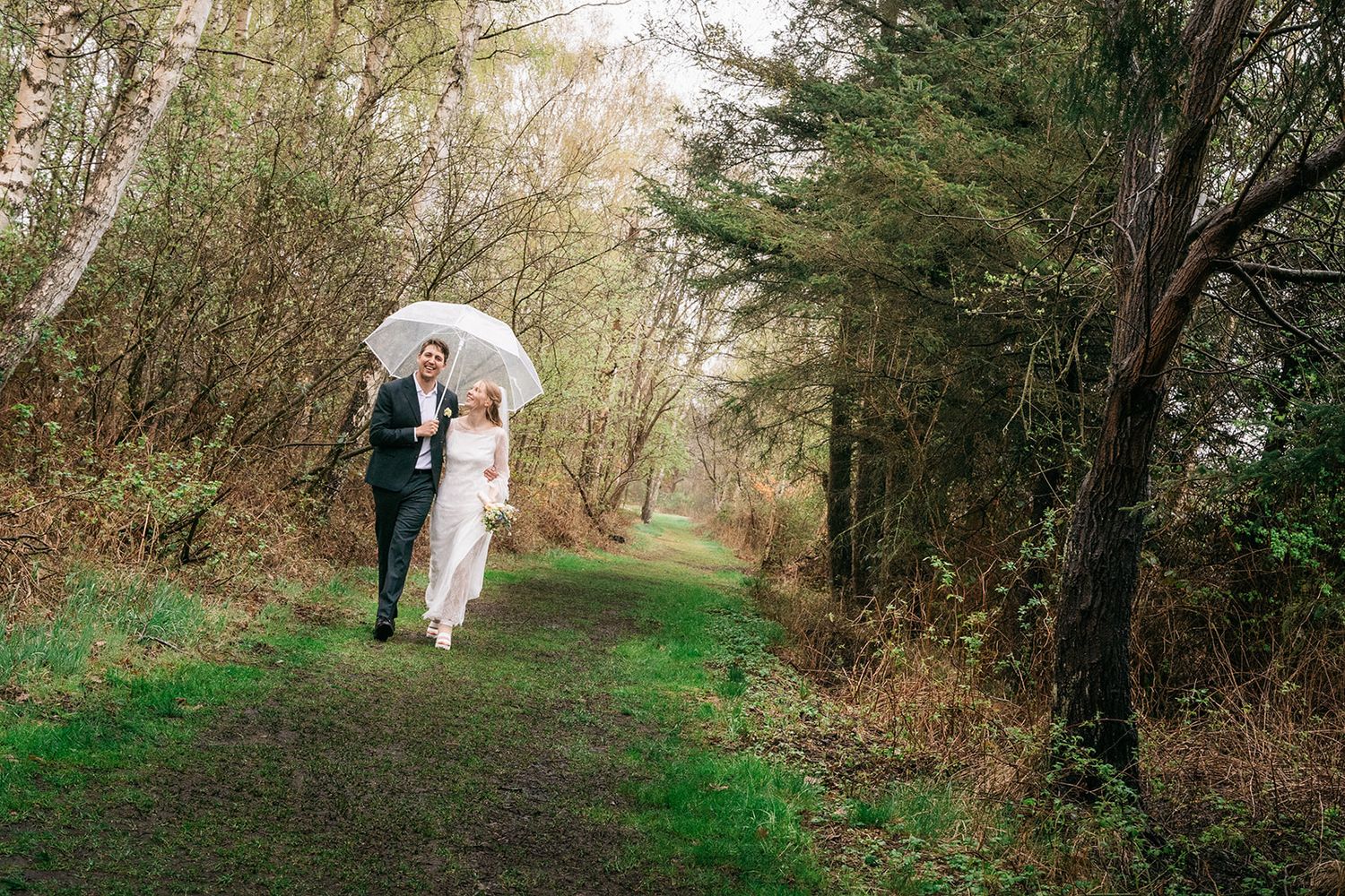 A couple shares an umbrella while walking along a scenic forest path on an overcast day.