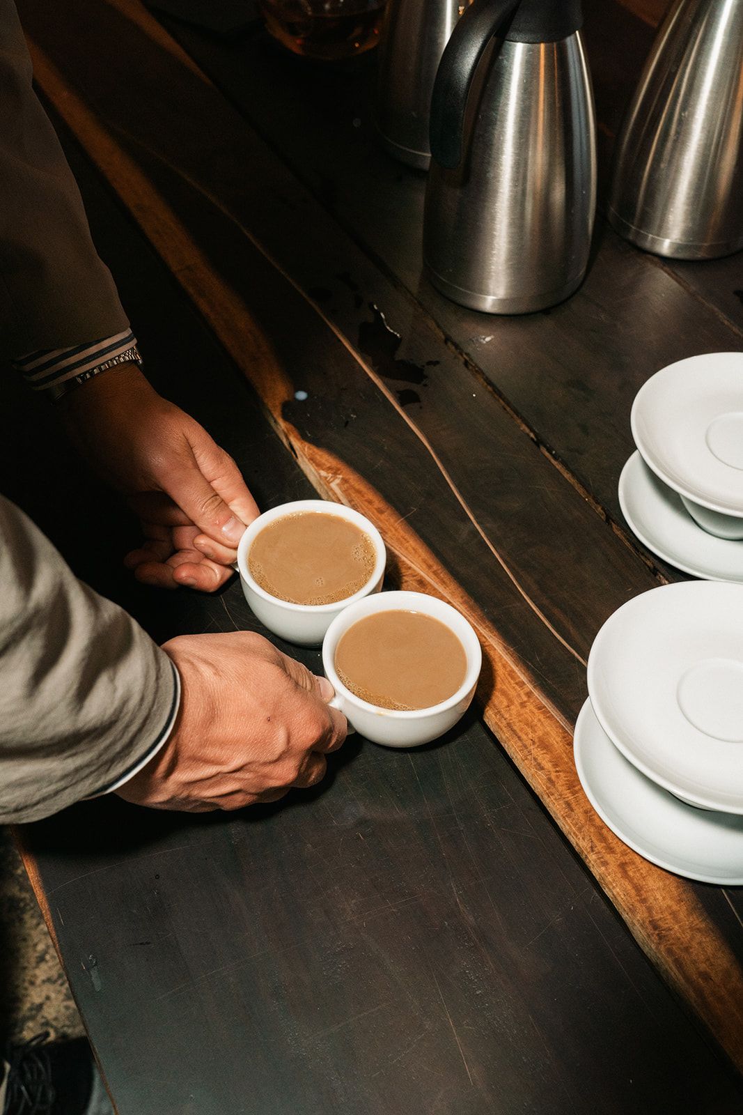 Two cups of creamy coffee or hot chocolate being served on a rustic wooden table with stainless steel thermos in background.