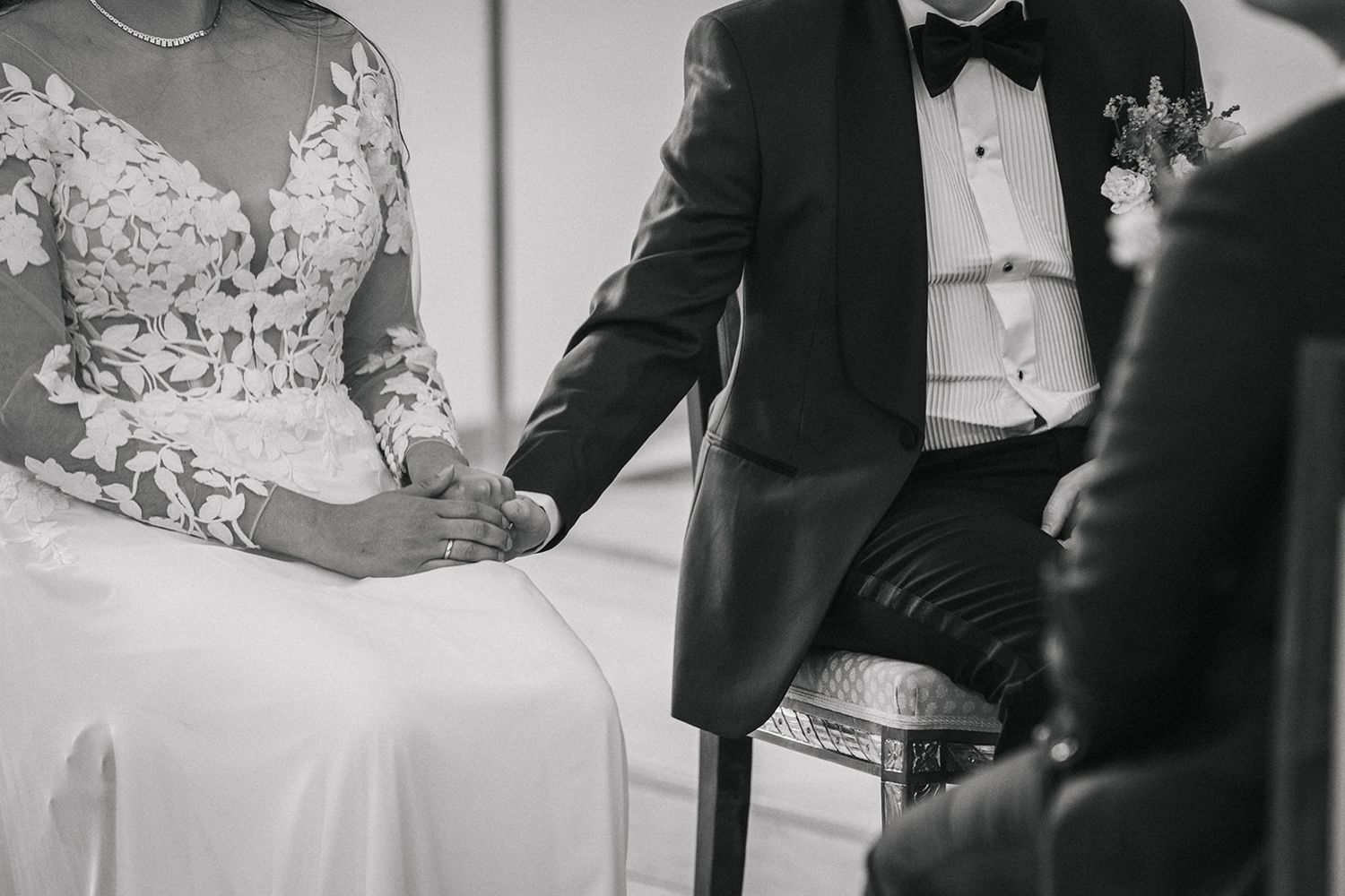 Black and white close-up image of a couple sitting together at their wedding, showing elegant dress and tuxedo details.