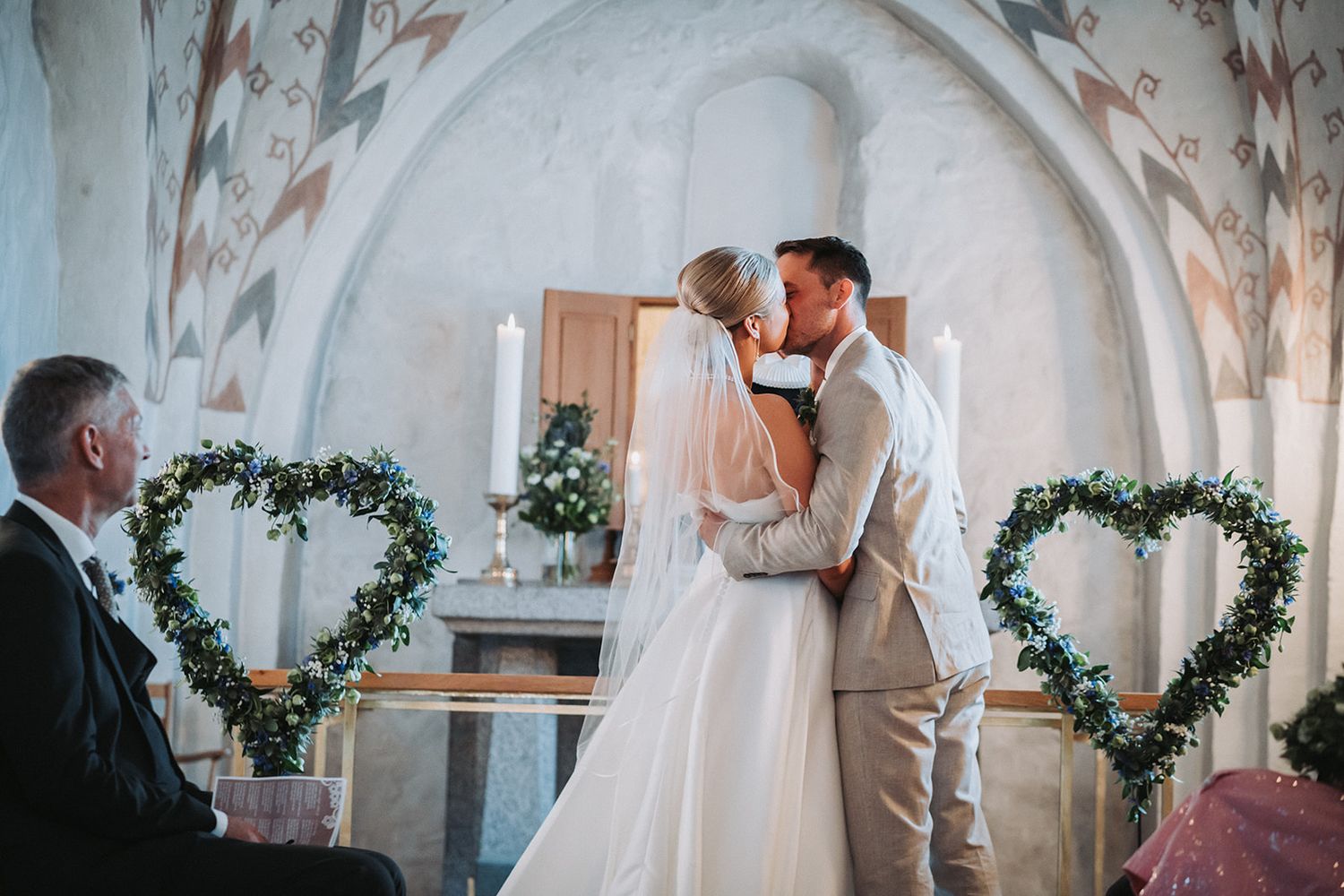 A couple shares their first kiss during an intimate wedding ceremony in a rustic chapel with heart-shaped greenery decor.
