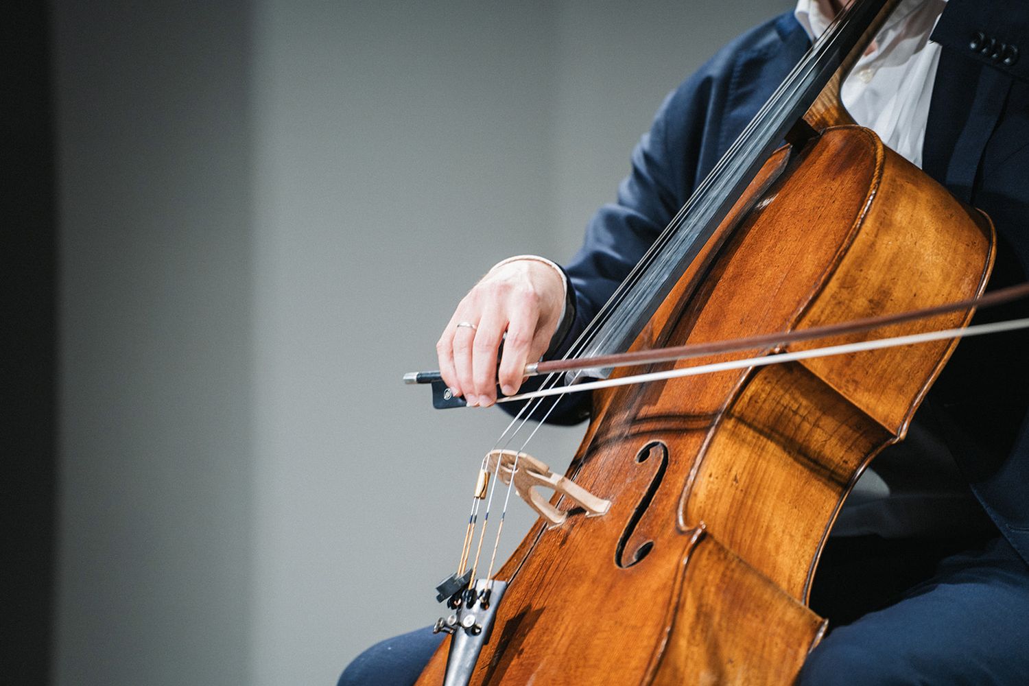 Close-up of musician playing a wooden cello with bow showing hands and instrument details against blurred background.