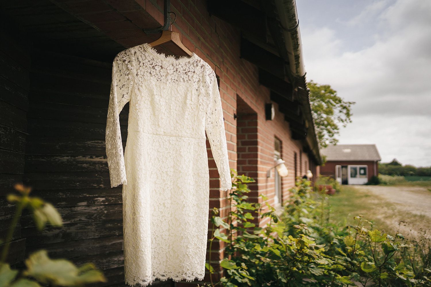 White lace wedding dress hanging on rustic barn wall with yellow wildflowers and countryside view in background.