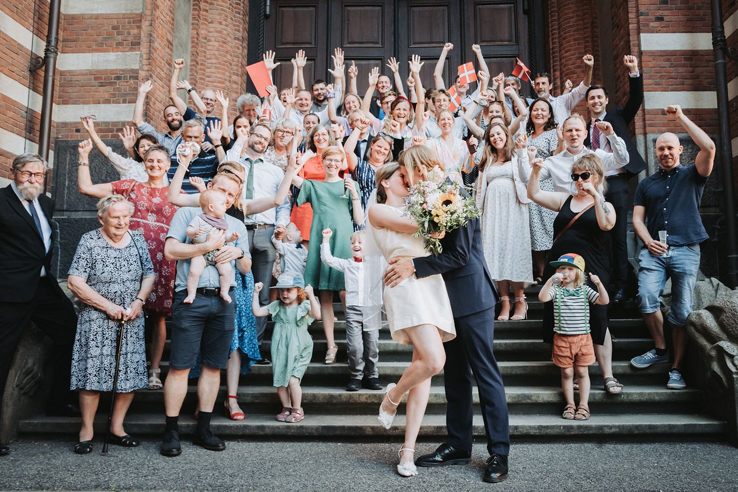 Large wedding celebration group photo on church steps with everyone cheering and raising their arms in celebration.