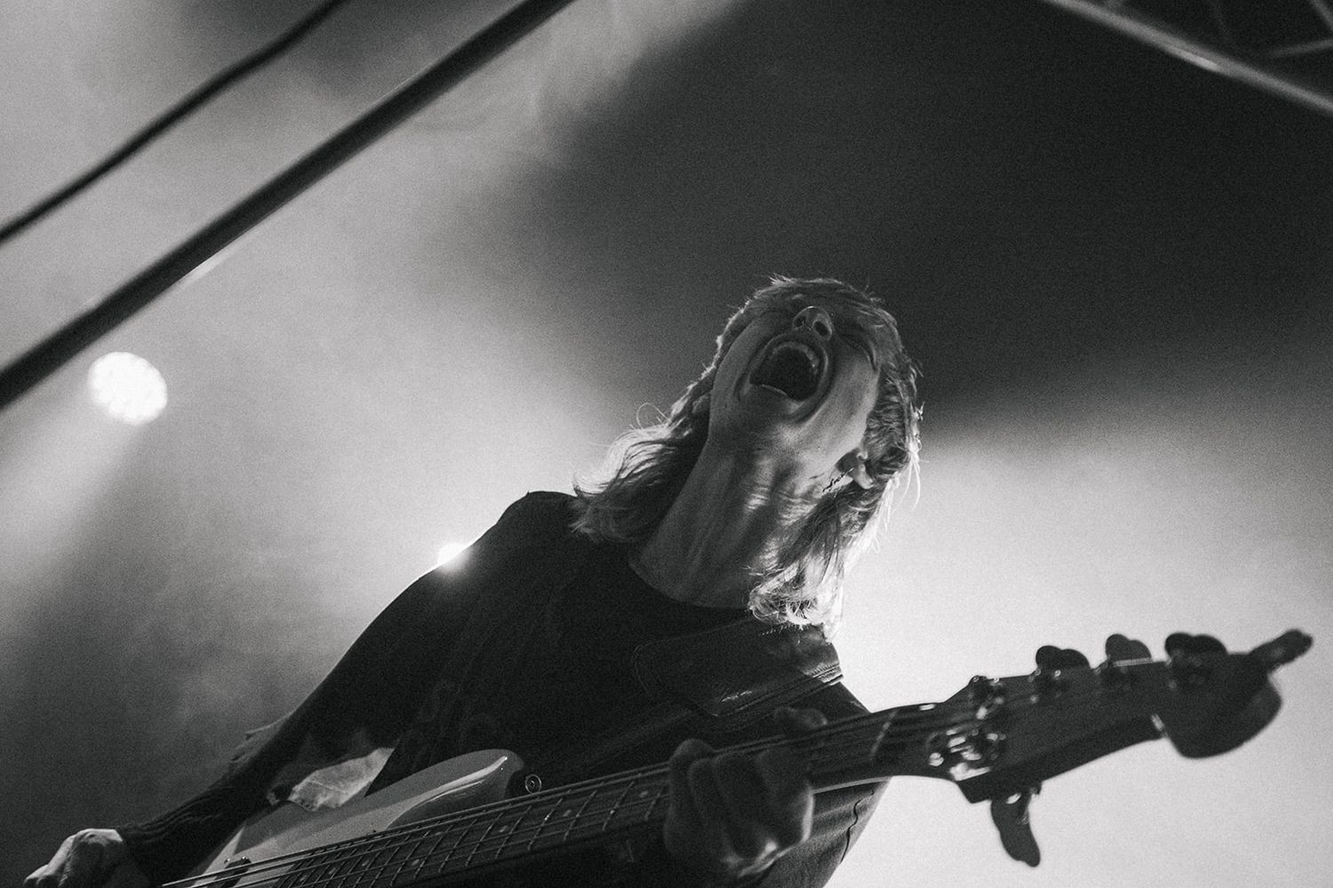 Black and white performance photo of a bassist playing on stage with dramatic lighting and long hair.