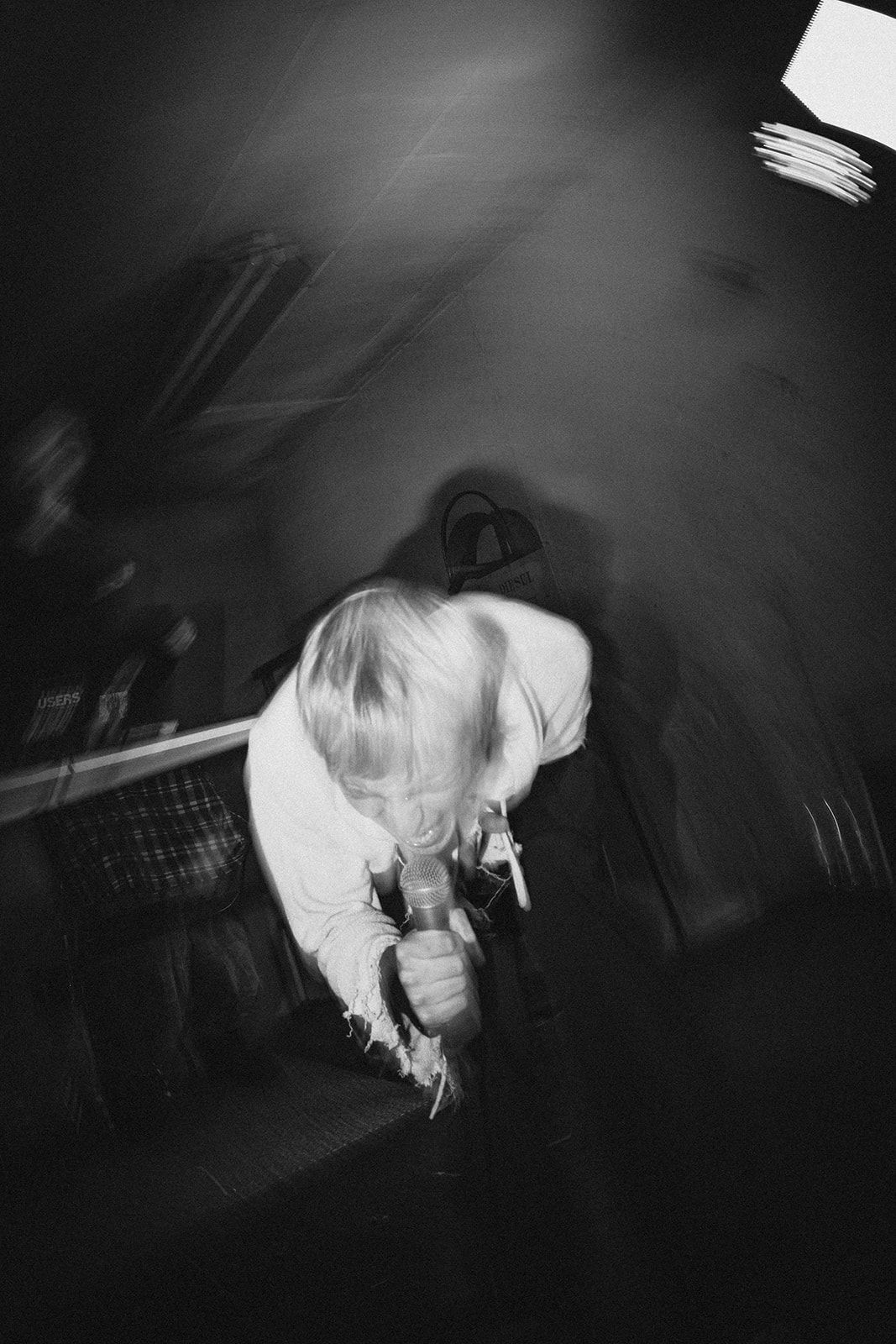 Black and white photo of someone with light hair leaning over a microphone in a dark performance setting.