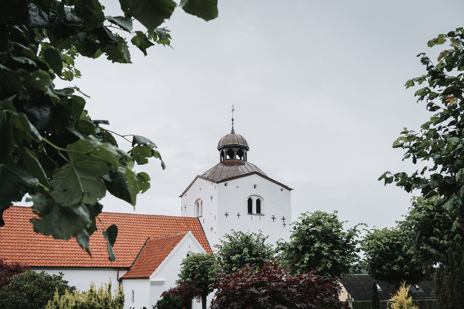 A traditional white church with a tower steeple rises above red-tiled roofs and green trees against an overcast sky.