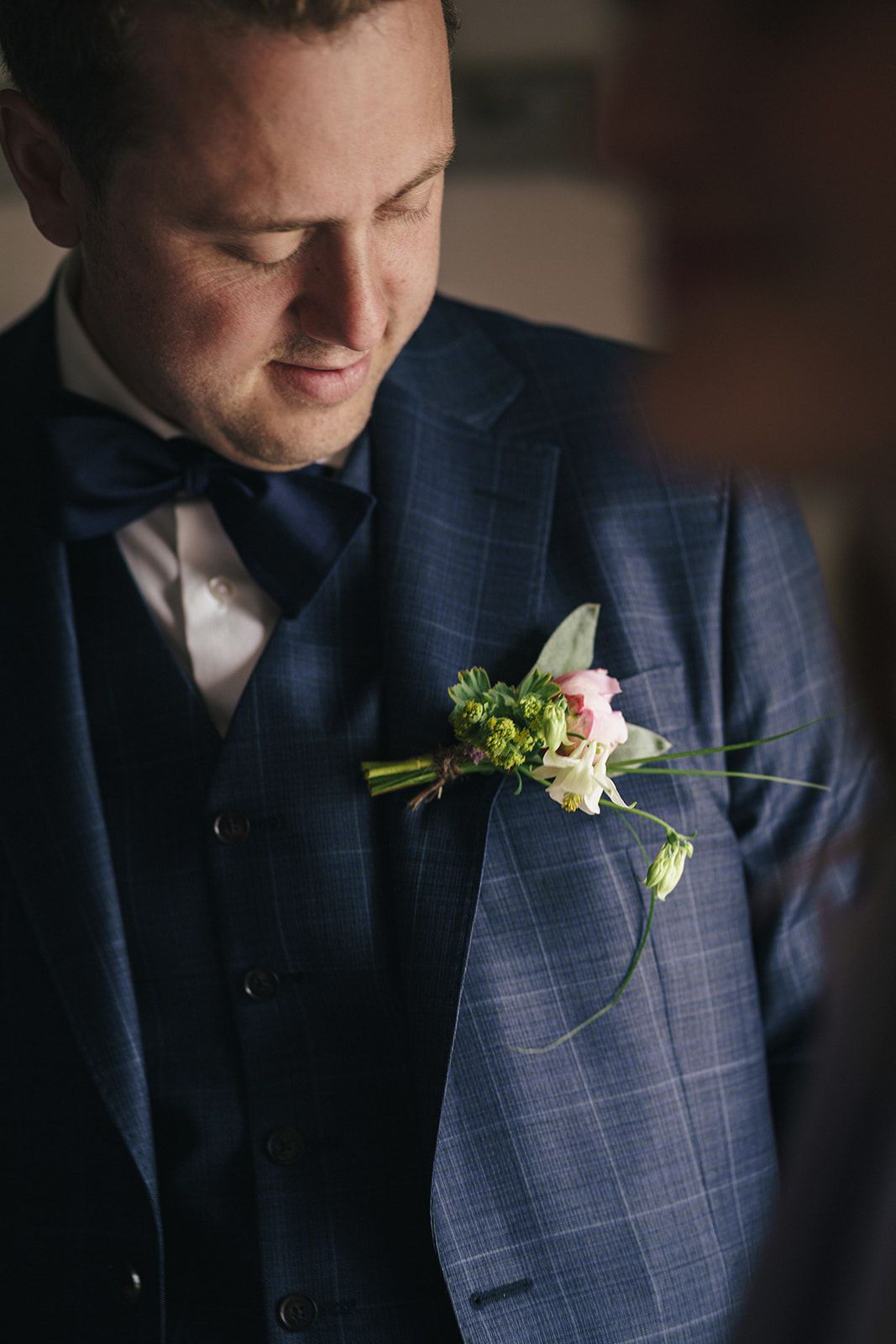 Close-up detail of groom wearing navy windowpane suit with delicate floral boutonniere in soft lighting.