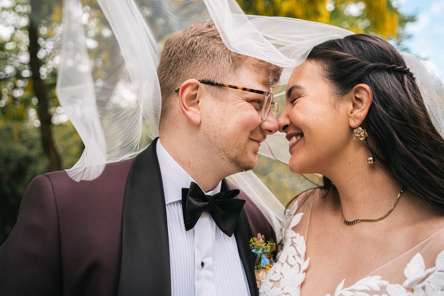 A happy couple shares an intimate moment outdoors with a flowing veil and autumn colors in the background.