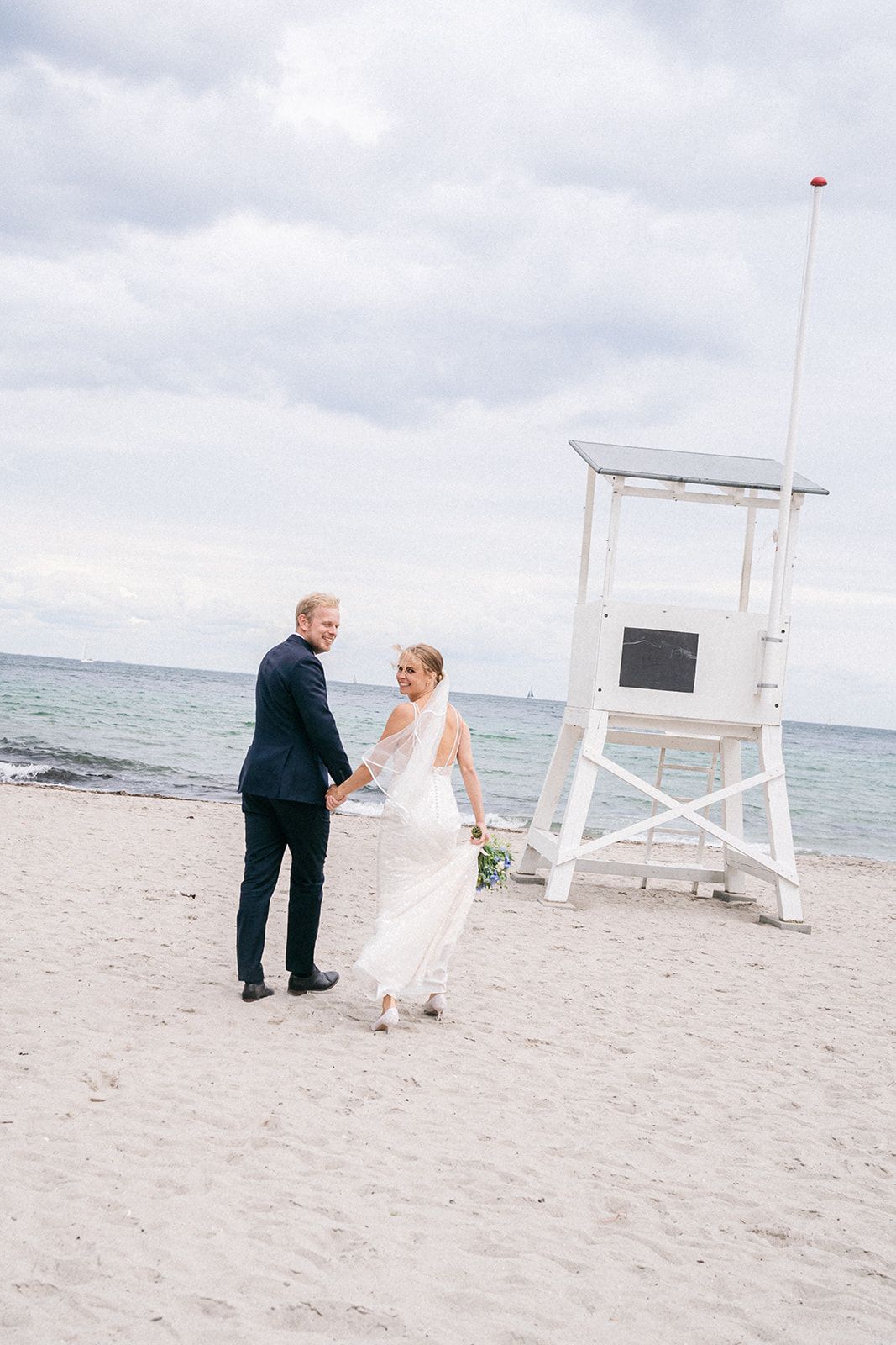 Wedding couple poses and walks along sandy beach with white lifeguard tower overlooking ocean on cloudy day.