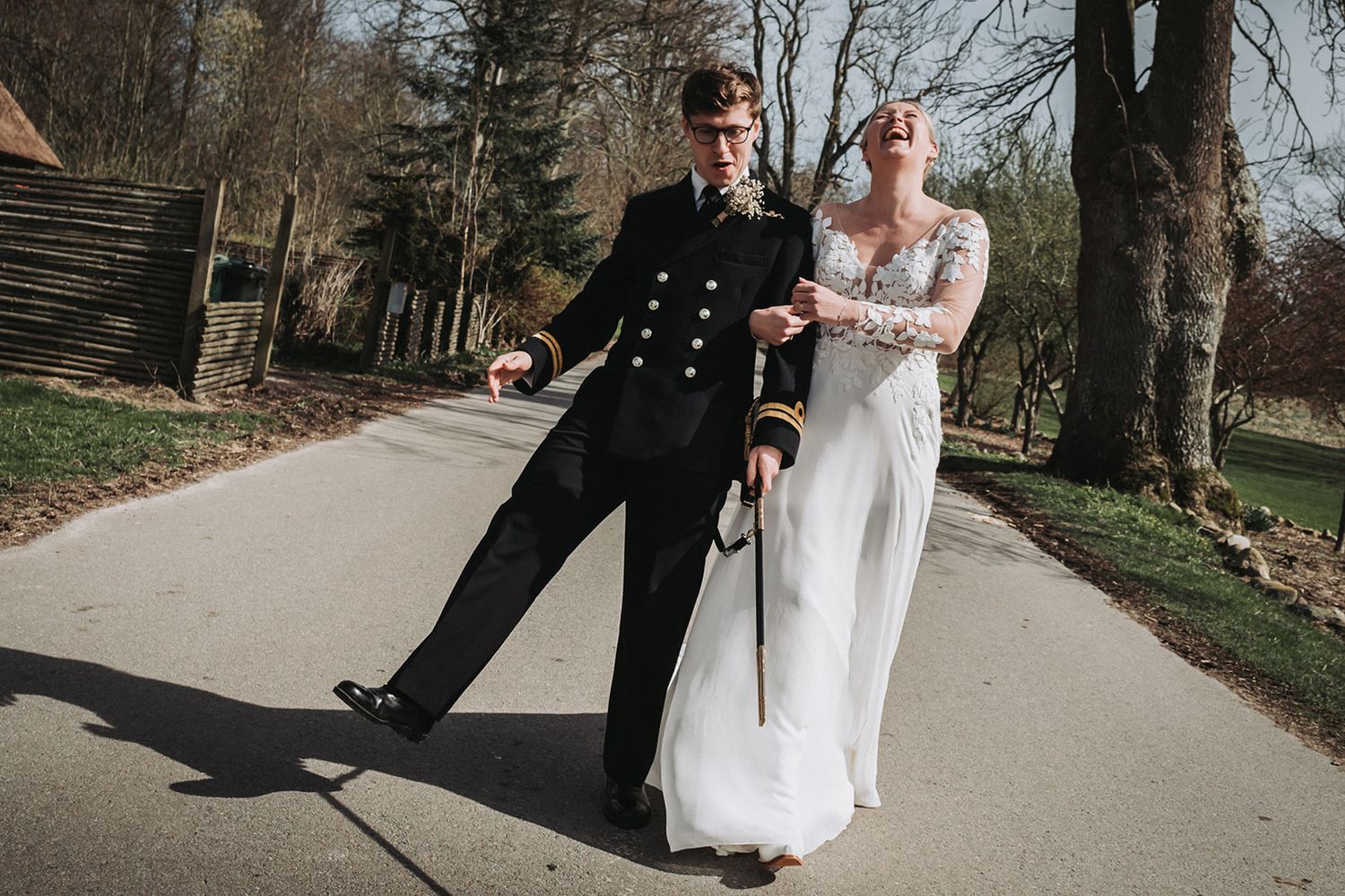 A couple walks together on a scenic path while taking creative and playful wedding photos in a park setting.