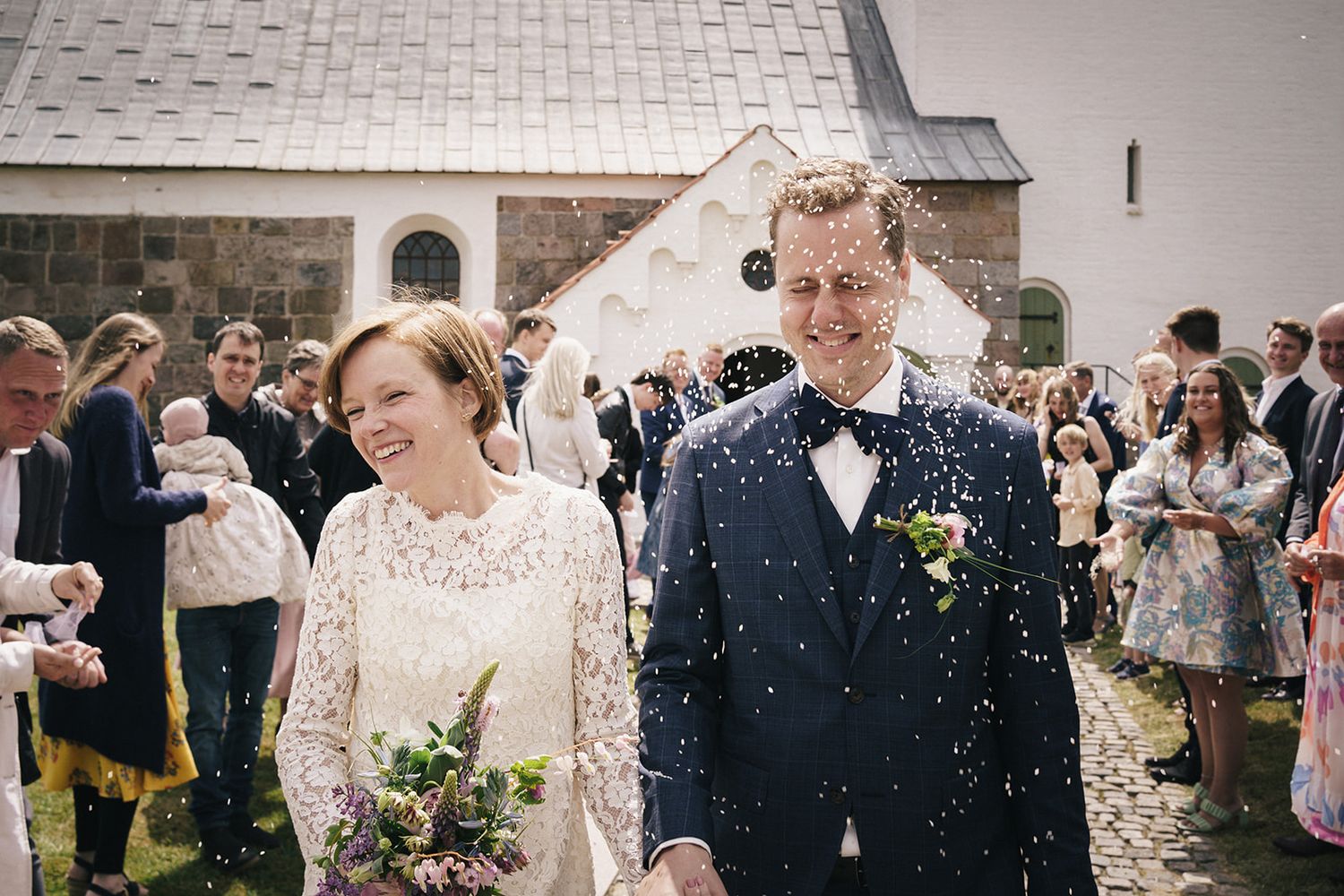 A joyful wedding couple exits a white church while guests shower them with confetti on their special day.