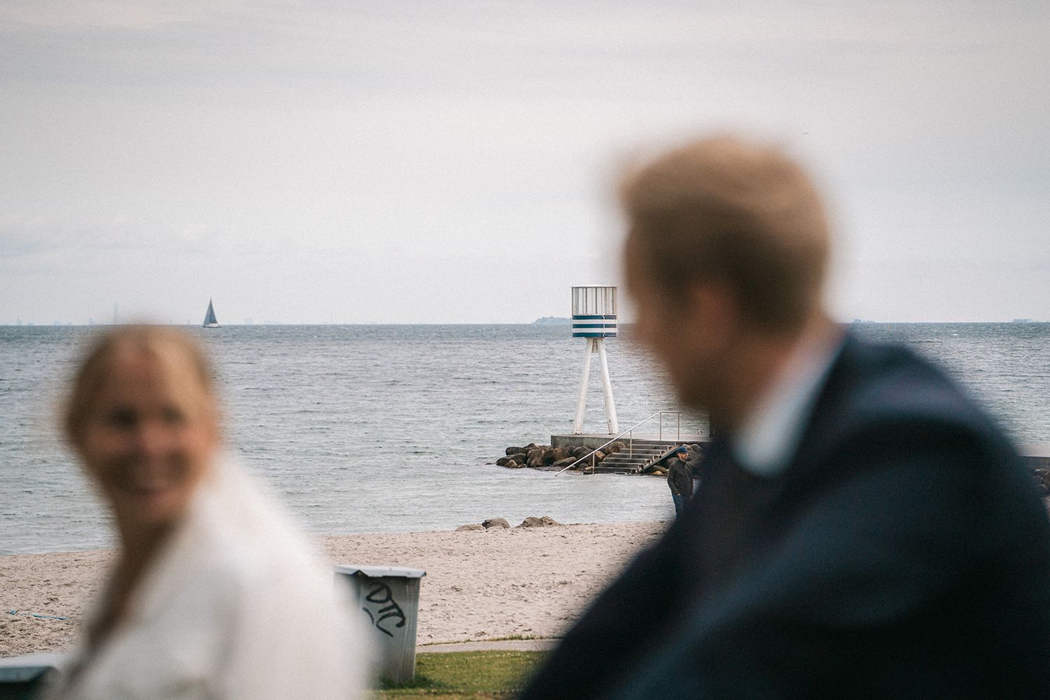 Two figures stand at a seaside location with a distant lifeguard tower and sailboat visible on the horizon.