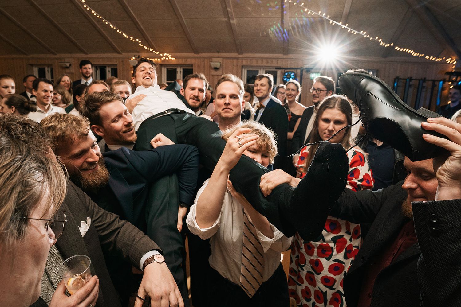 A crowd of energetic guests dance together and celebrate under string lights at a lively indoor event reception.