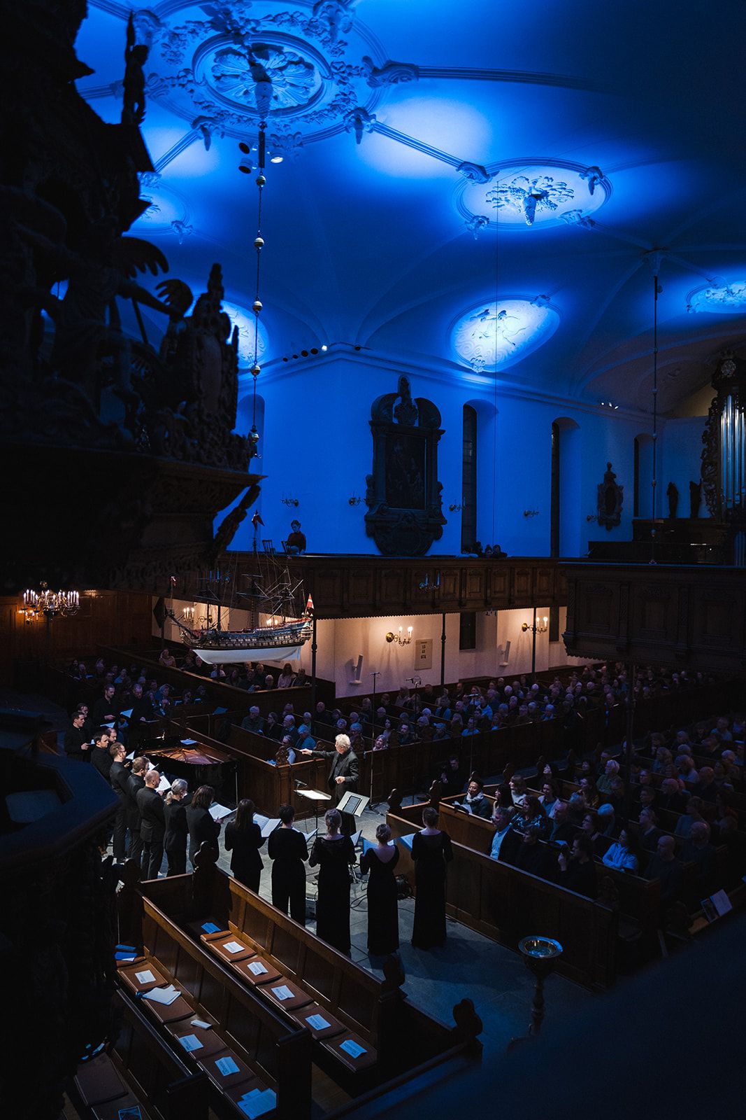 A choir performs in a dimly lit cathedral with dramatic blue lighting illuminating ornate ceiling fixtures and architecture.