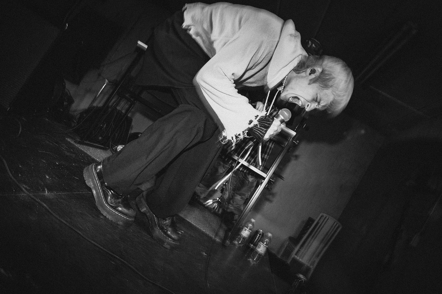 Black and white image of a performer on stage playing an electric guitar with intense energy while bending down.
