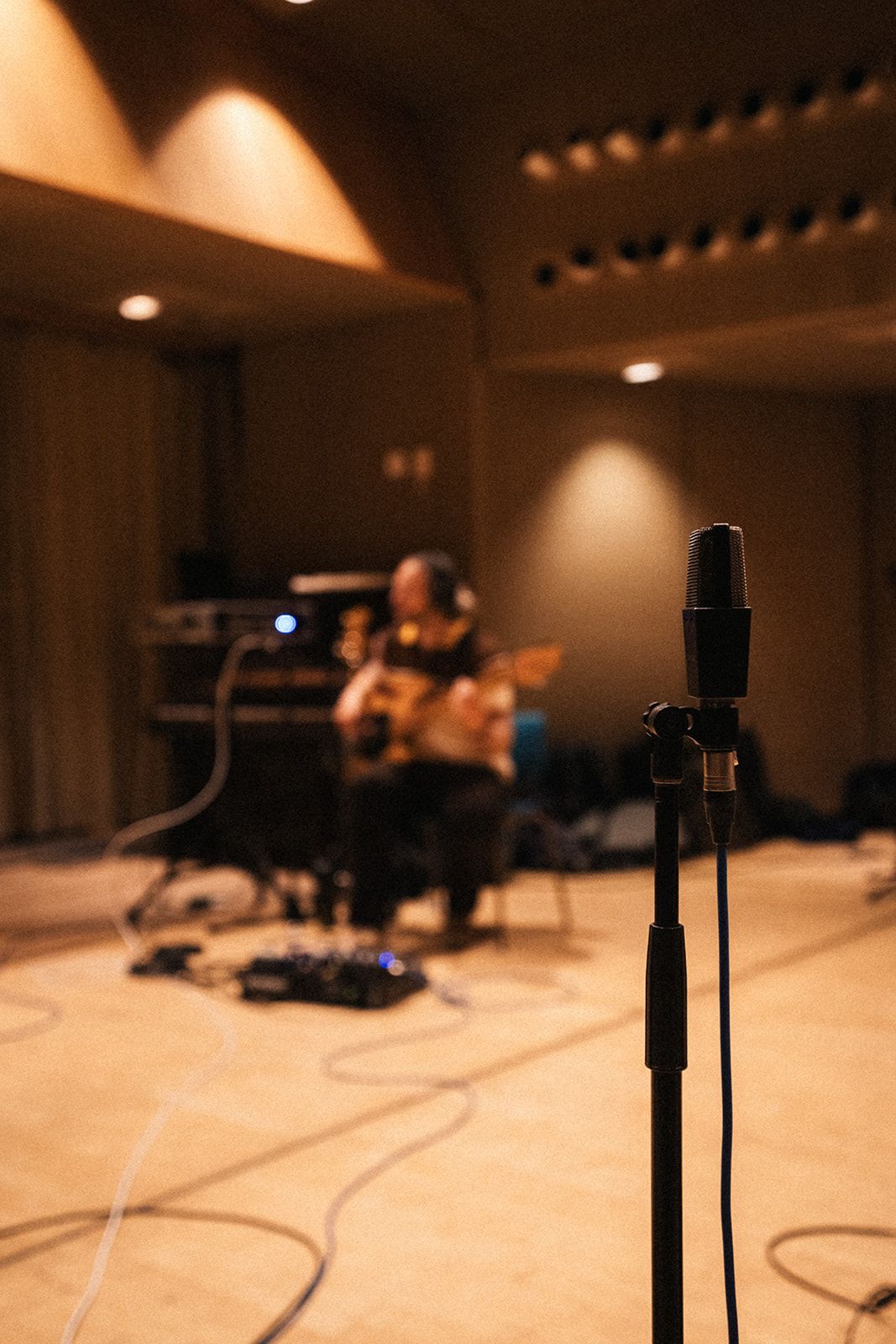 A microphone stand in focus with blurred recording studio and guitarist in the background during a session.