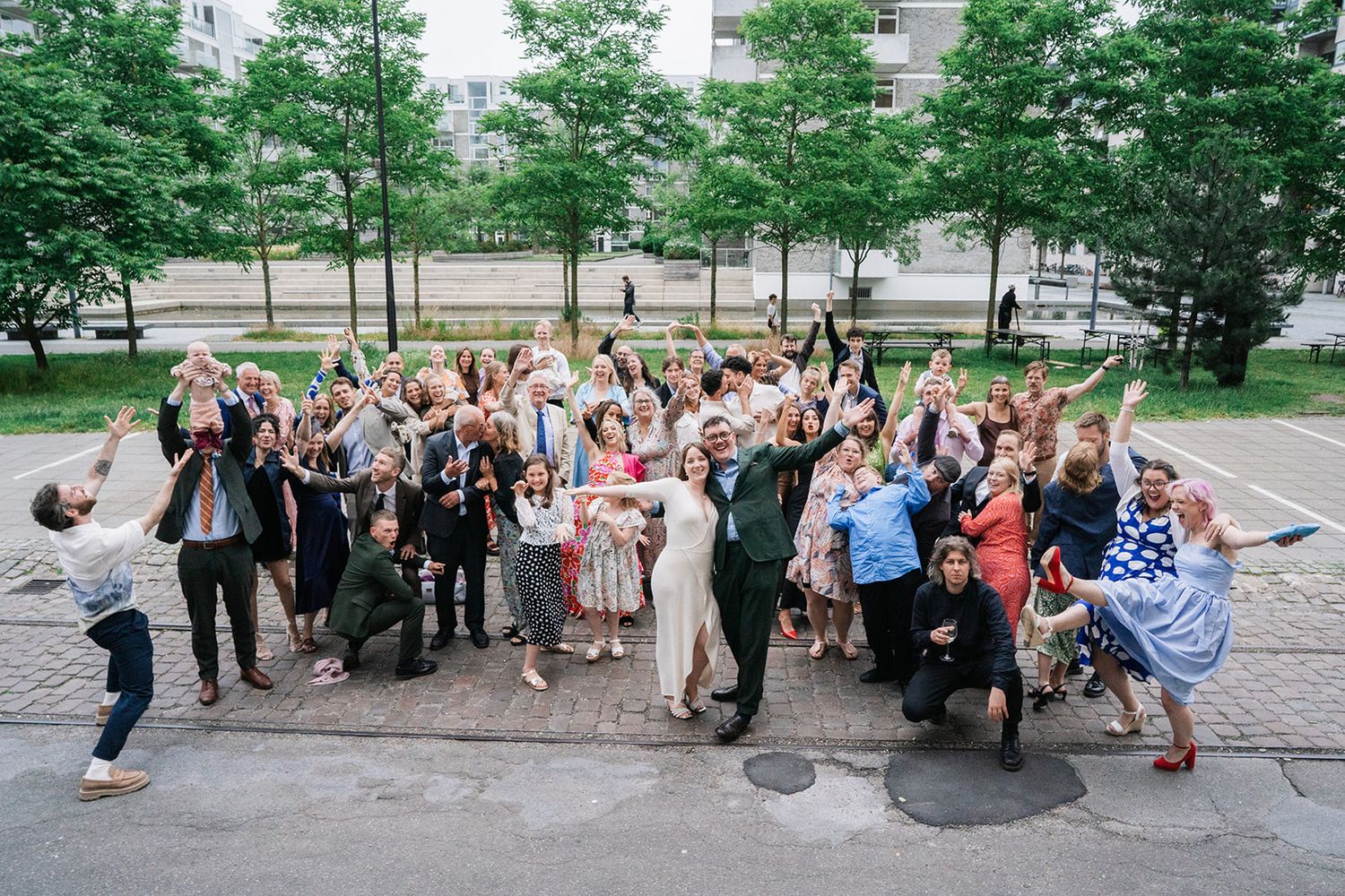 Large group of wedding guests posing together outdoors with raised arms in celebratory gestures on stone steps.