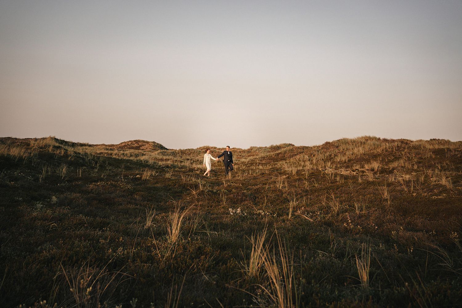 Two figures stand atop a grassy hillside against a moody sunset sky in a romantic landscape setting.