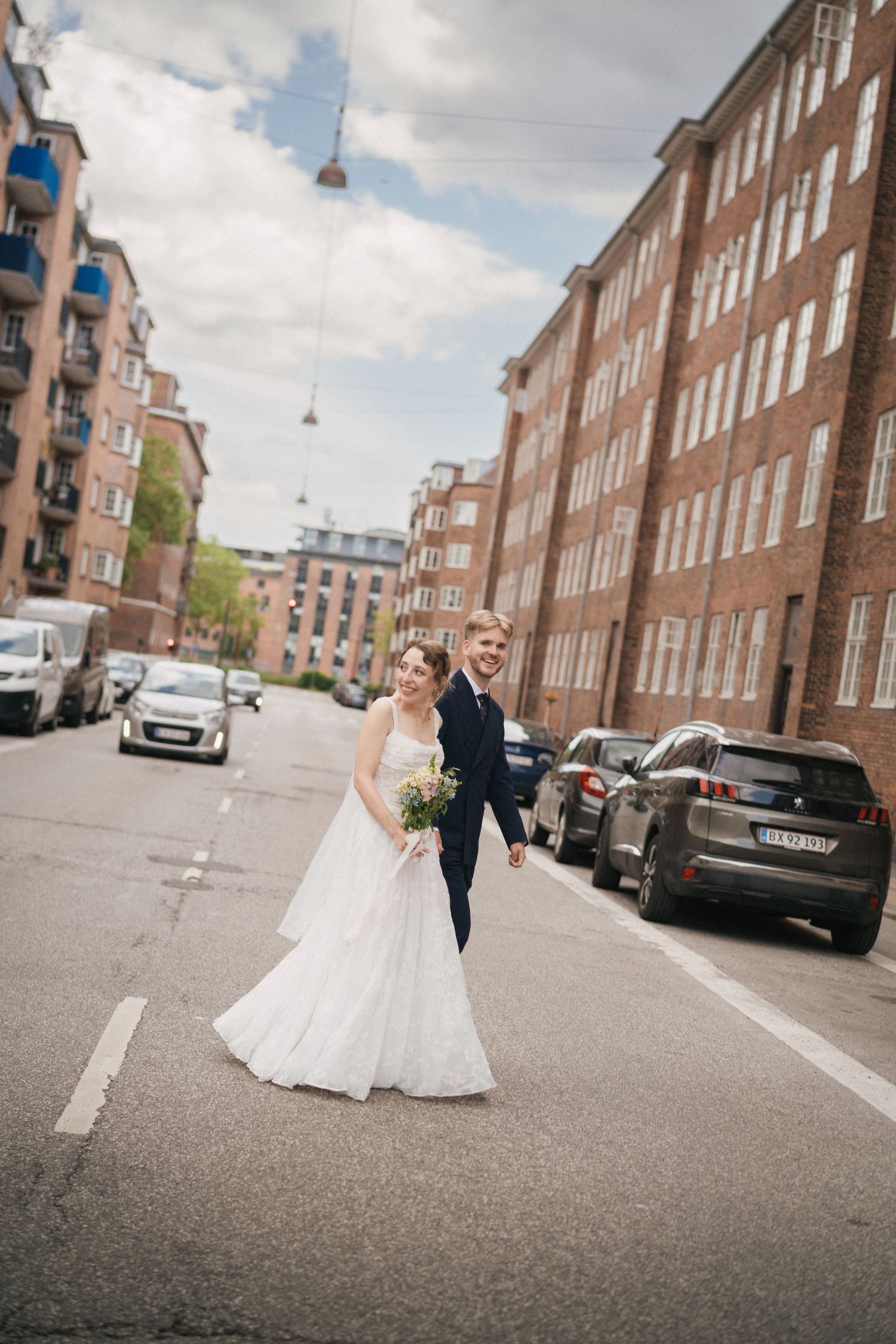 Bride and groom walking hand in hand on a city street, surrounded by brick buildings and parked cars.