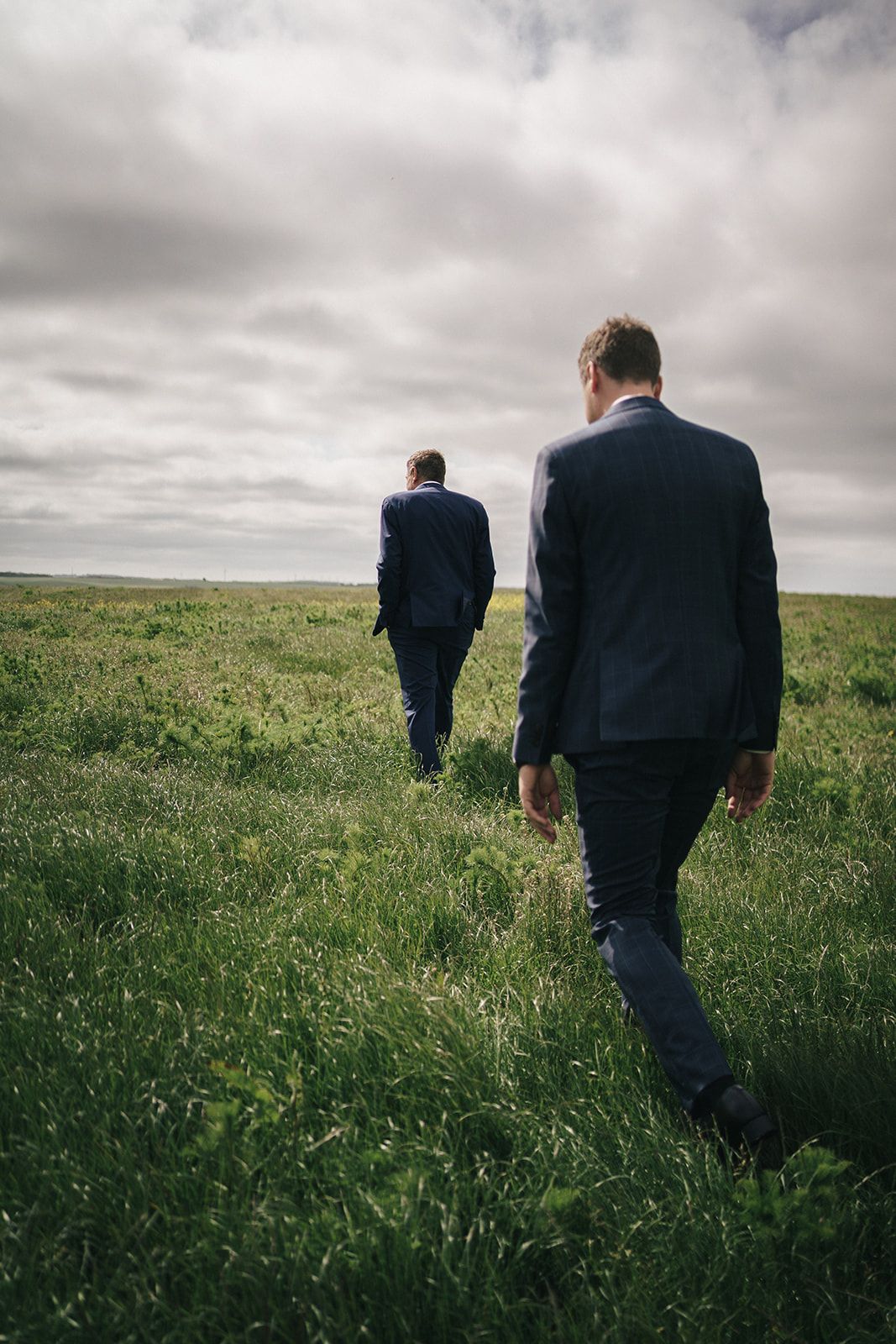 Two men in dark suits walk through a green field under moody clouds on a somber day.