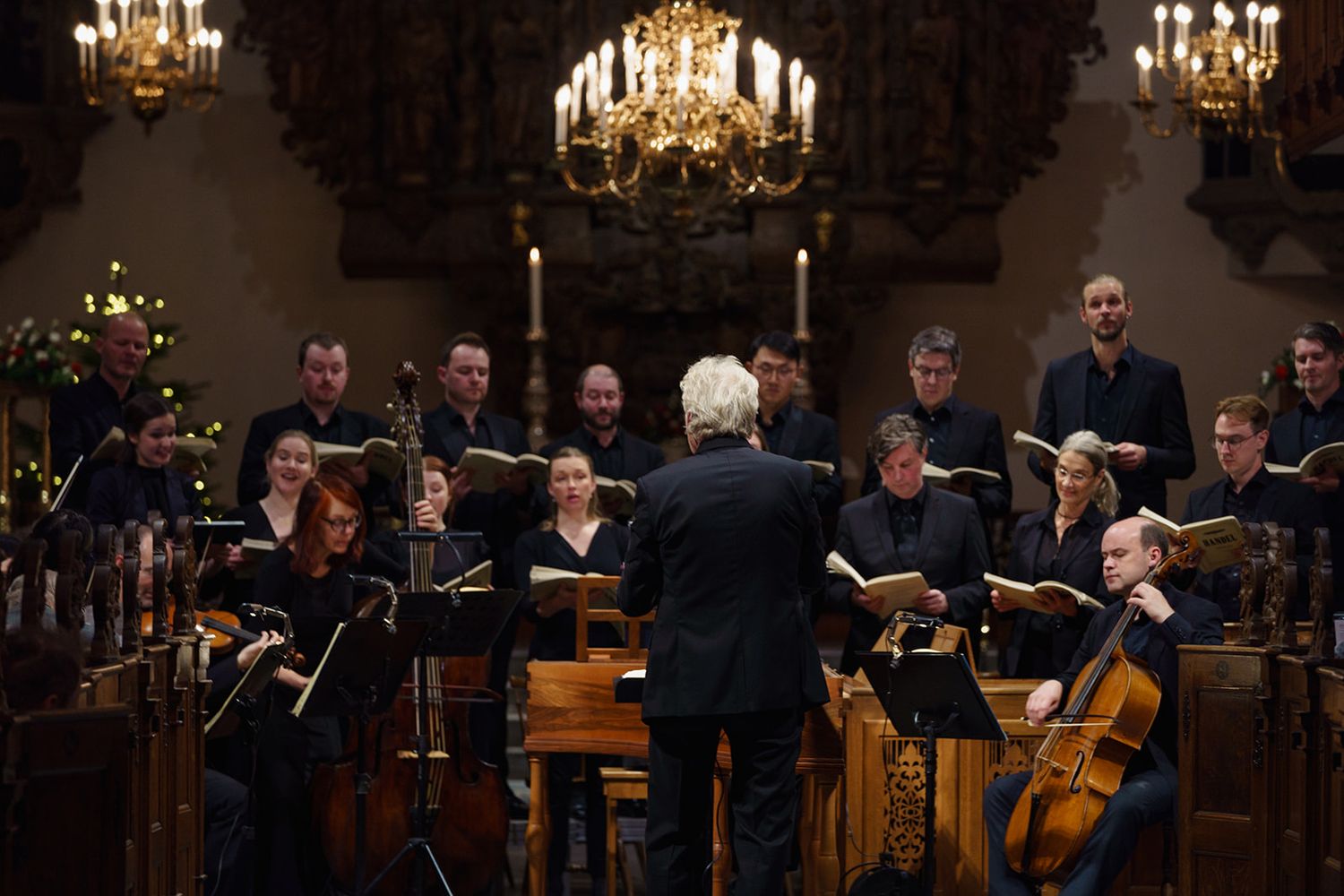 Orchestra and conductor perform in elegant concert hall illuminated by ornate chandeliers during classical music performance.