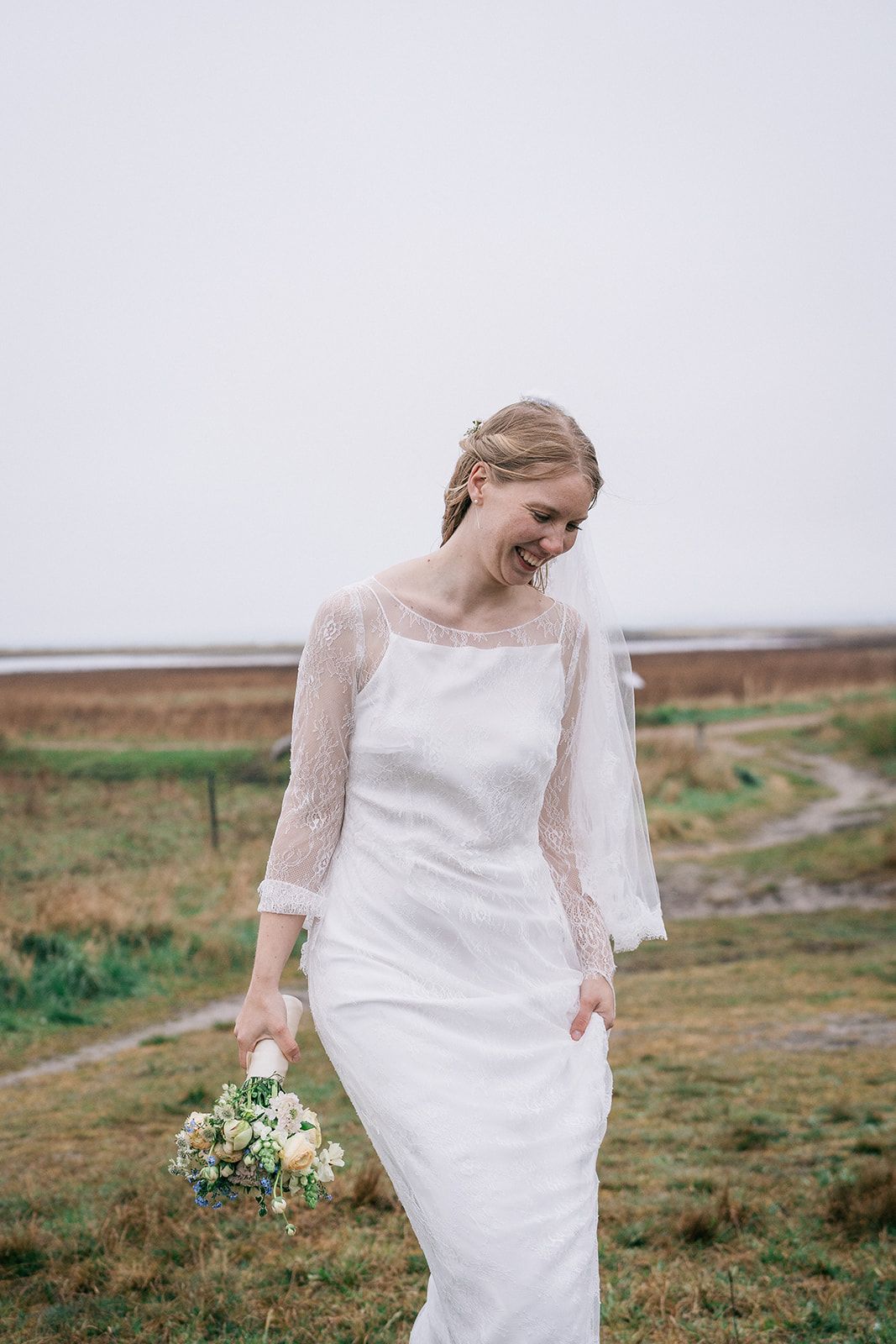 A bride in a white lace wedding dress walks through a green meadow holding a small white floral bouquet.