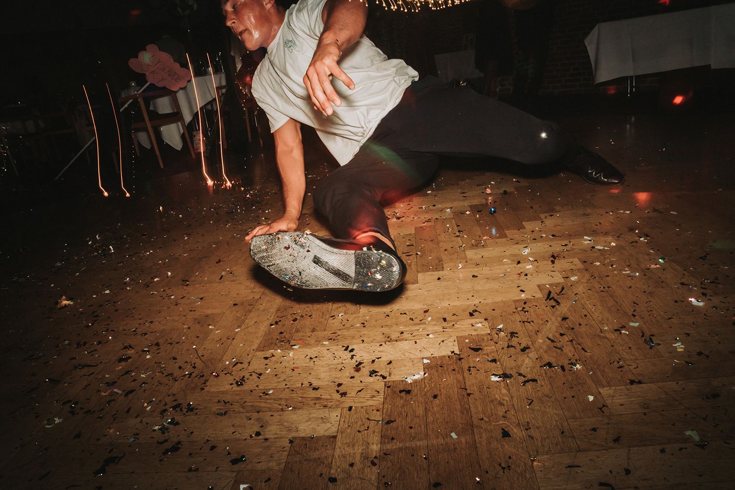 A person performs an energetic skateboard trick on a scattered debris-covered wooden floor at night.