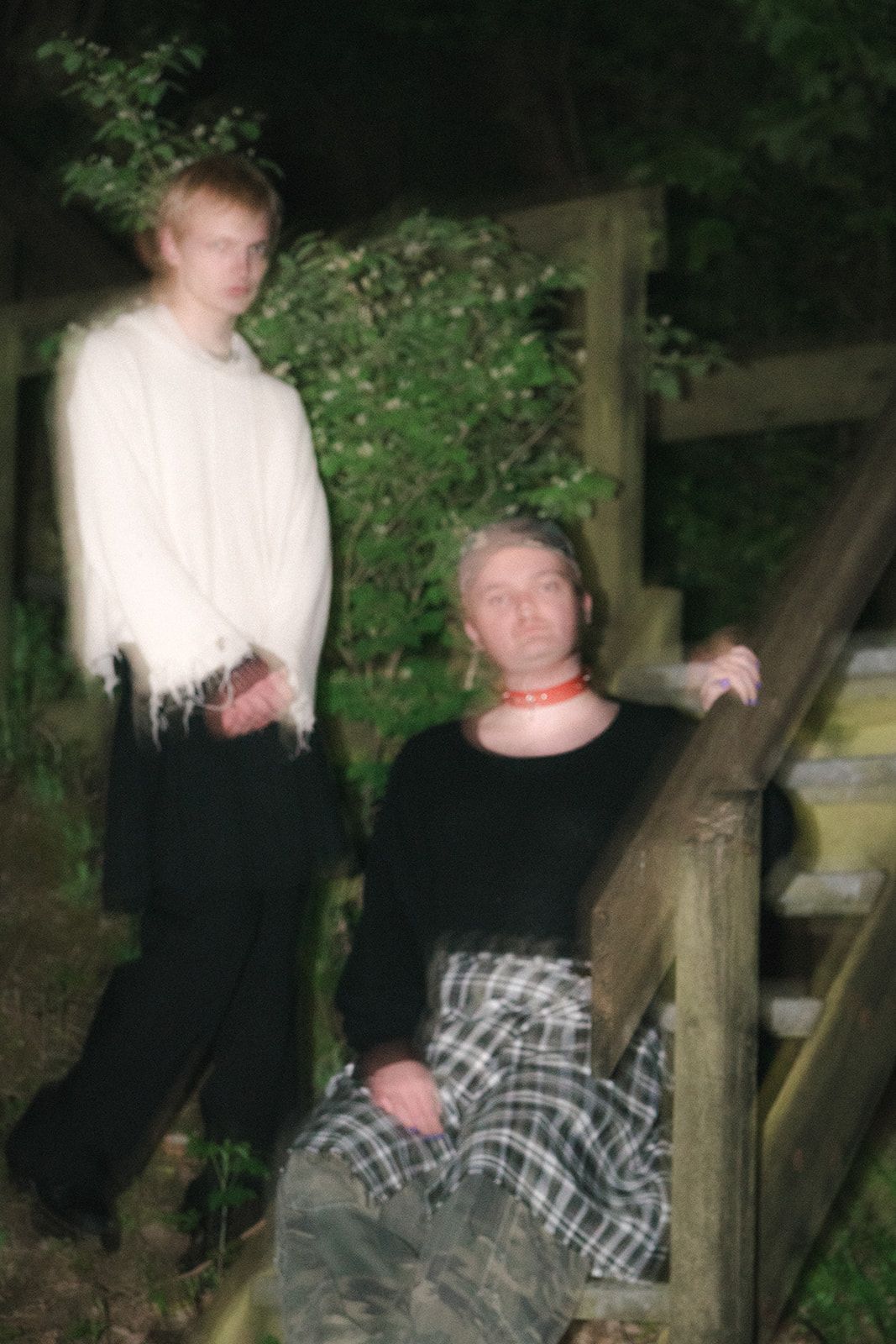 Two people pose at night on wooden steps outdoors, one standing in white and one sitting in black with plaid skirt.