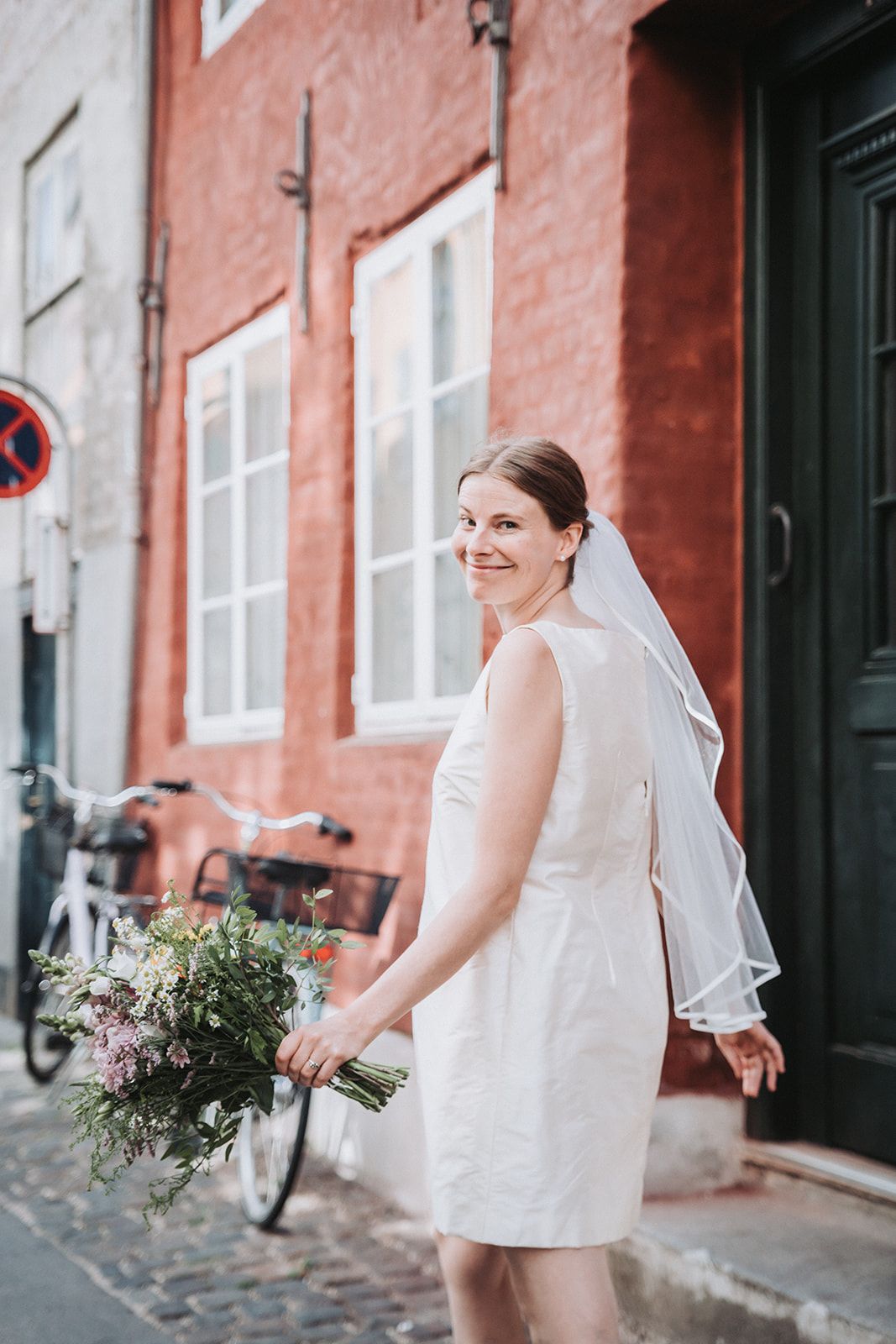 Smiling bride in a short white dress with veil holds wildflower bouquet outside brick building with bike in background.