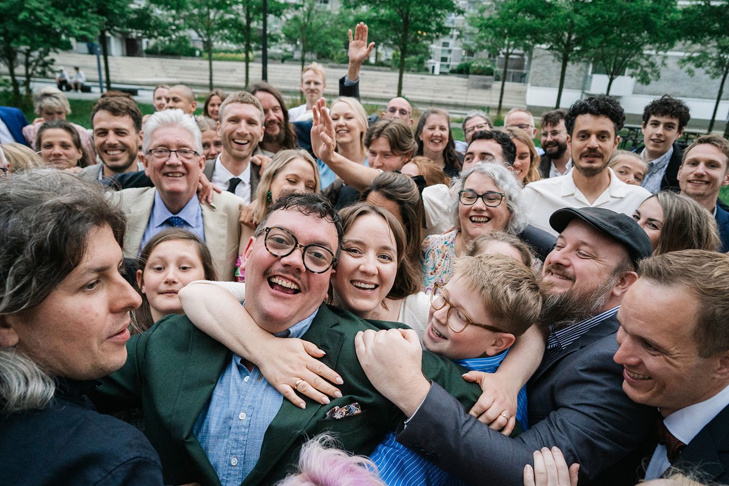 A cheerful crowd of people hugging and celebrating together outdoors with trees in the background.