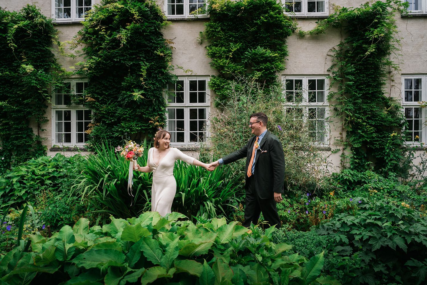 Wedding couple embraces in romantic garden setting with historic stone building and lush greenery in the background.