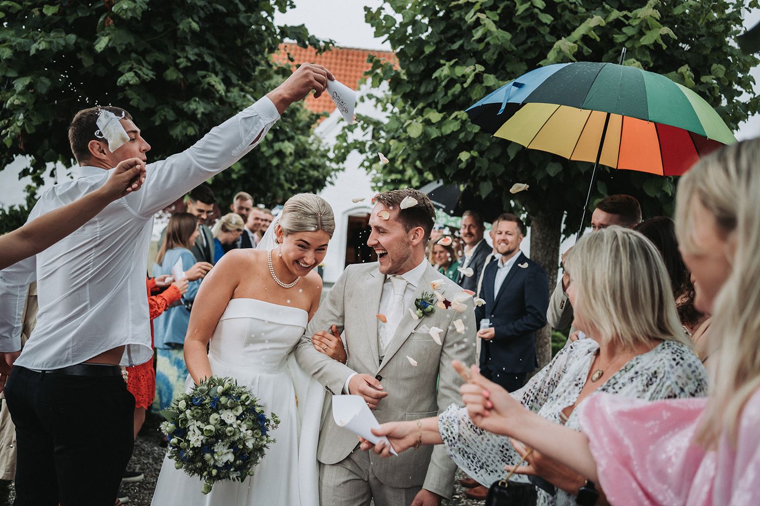 Wedding party celebrating under a colorful umbrella with guests tossing confetti in an outdoor garden setting.