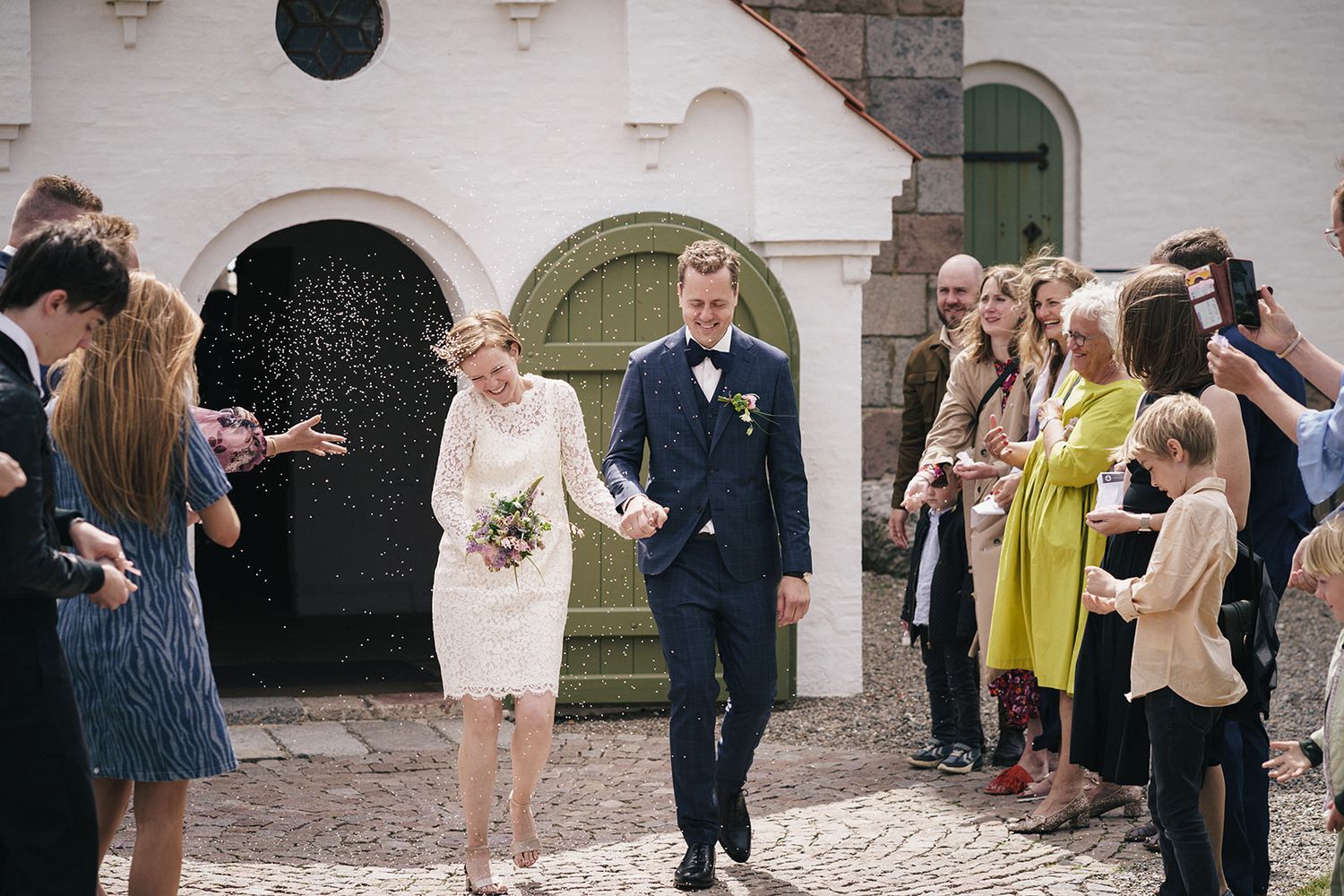 Wedding guests lined up outside white church entrance throwing flower petals during celebration.