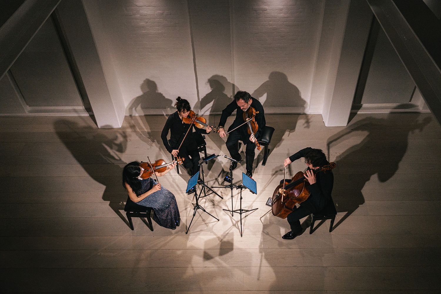 Aerial view of musicians practicing in a circle formation with dramatic shadows cast on bright white floor.