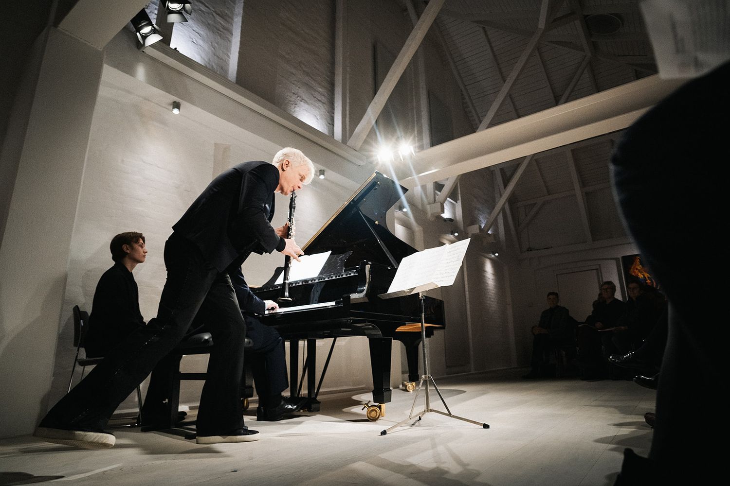 Musicians perform at a piano in a dimly lit concert space with dramatic lighting and high ceilings.