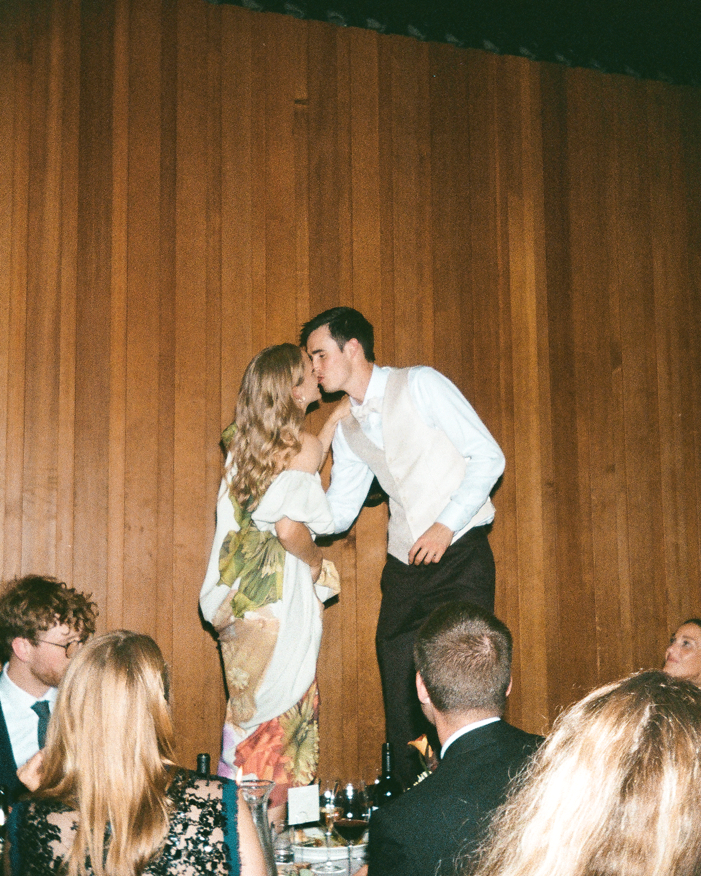 Couple sharing a romantic kiss at a formal event, standing above seated guests near a wooden panel wall.