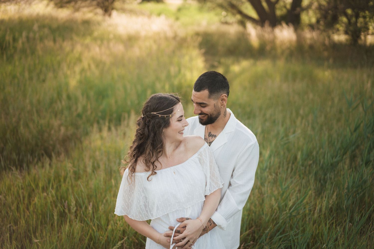 Couple in white outfits embracing in a sunlit green field during a romantic maternity photo session.