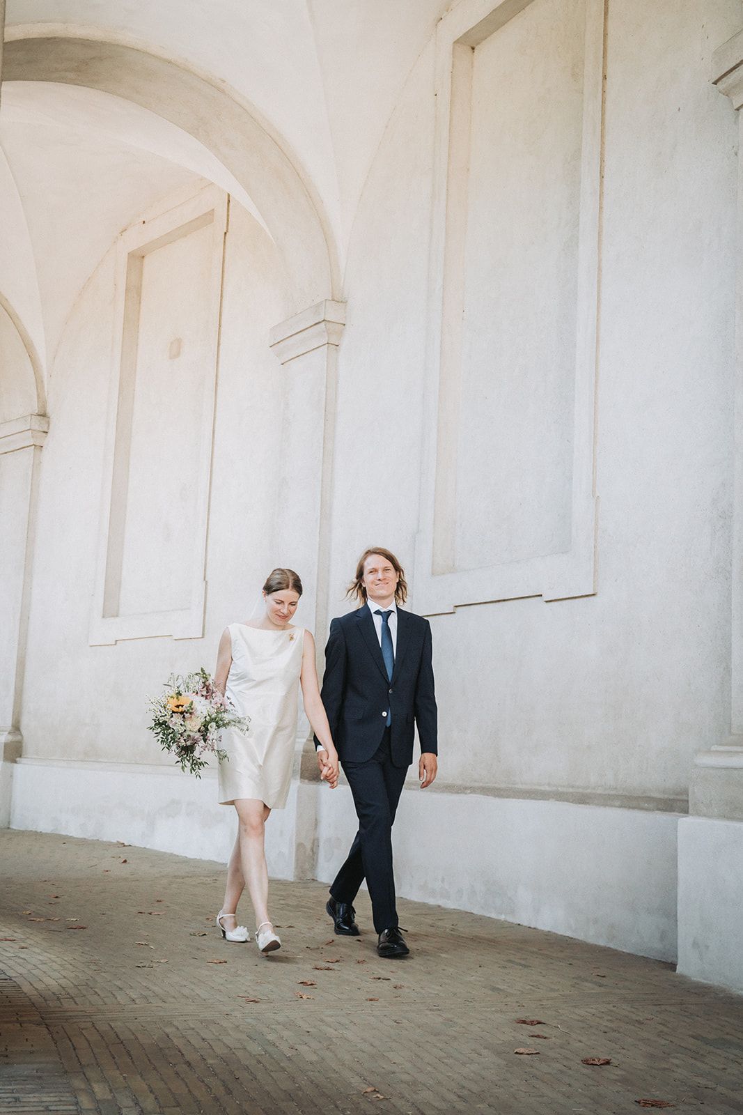 Elegant couple strolling through white arched hallway during wedding photoshoot.