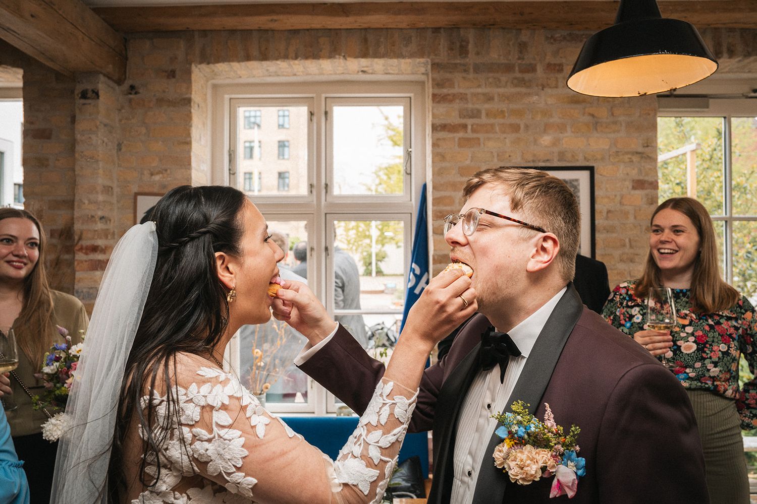 A couple in wedding attire playfully feed each other cake during a wedding reception in a rustic brick venue.