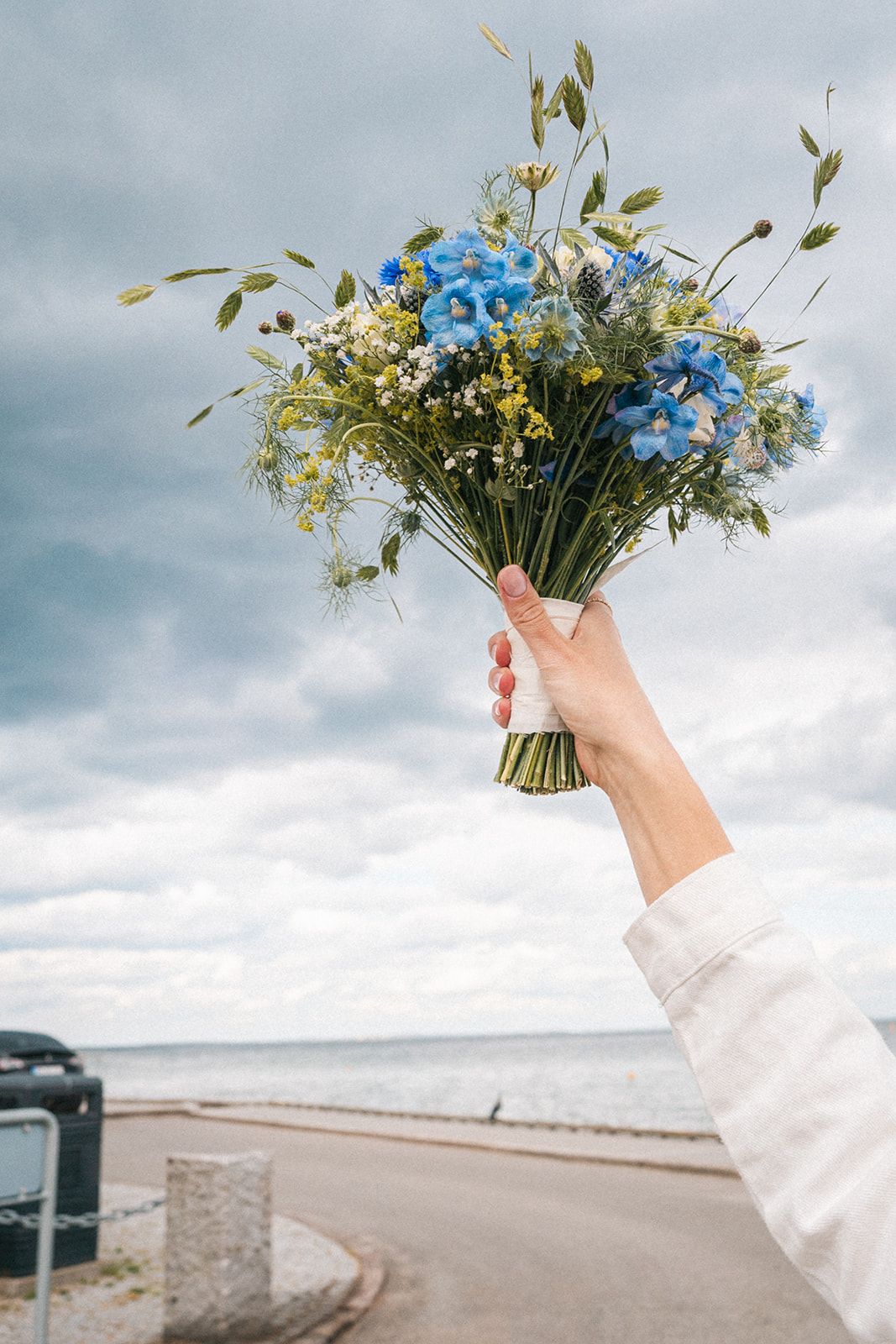 Hand holding up a beautiful wildflower bouquet with blue flowers and greenery against a cloudy sky at the beach.
